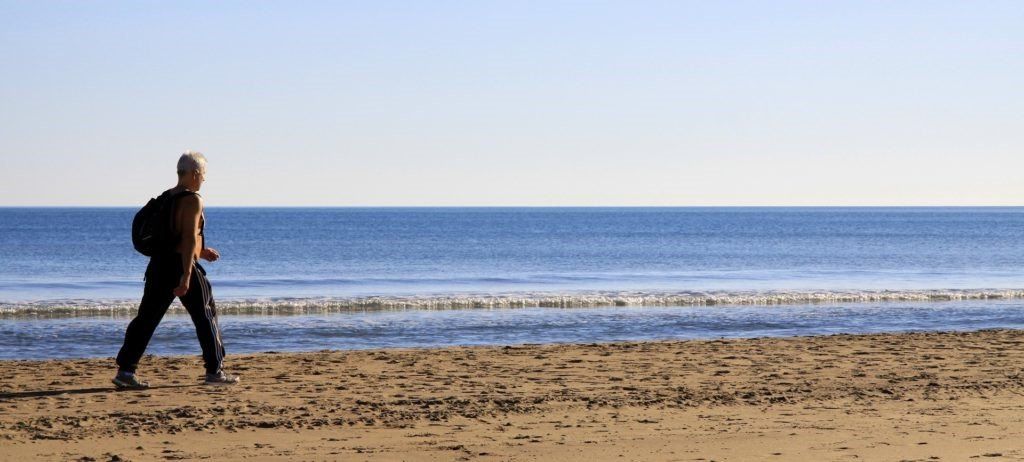 homem maduro caminhando por uma praia deserta. ele vesta calças pretas, mochila preta e camisa bege