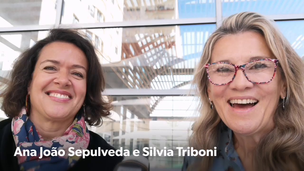 Maturis posam para foto juntas, ambas sorriem. A mulher da esquerda é morena, e tem os cabelos curtos. A mulher da direita é loira e usa óculos. Ambas sorriem.