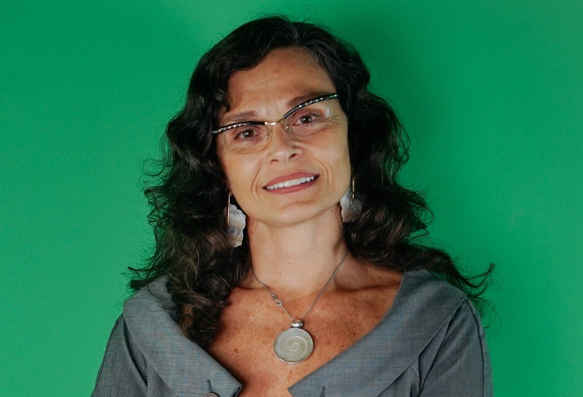 mulher de óculos de grau, cabelos longos ondulados, brincos e colar grandes, sorri para foto
