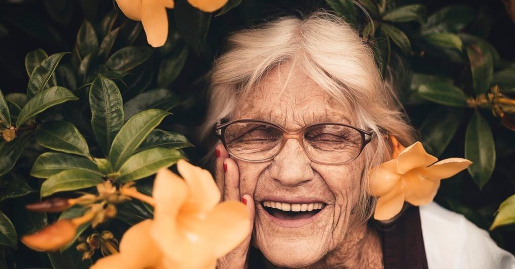 maturi sorri em meio à flores de cor laranja. ela usa óculos, está de olhos fechados e sorrindo enquanto apoia a mão no rosto. suas unhas são vermelhas e ela aparenta felicidade