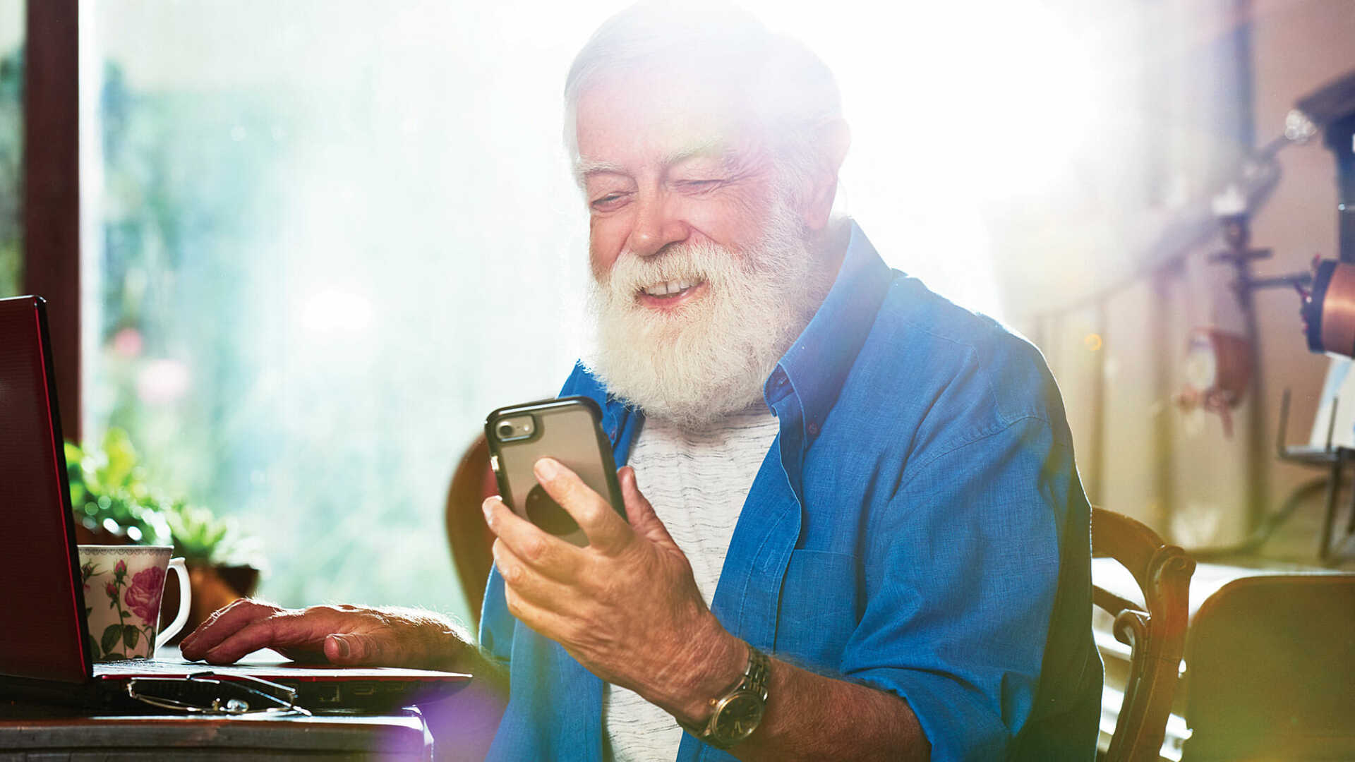 maturi olha para o celular e sorri demonstrando satisfação. ele veste uma camisa jeans e tem uma longa barba branca.