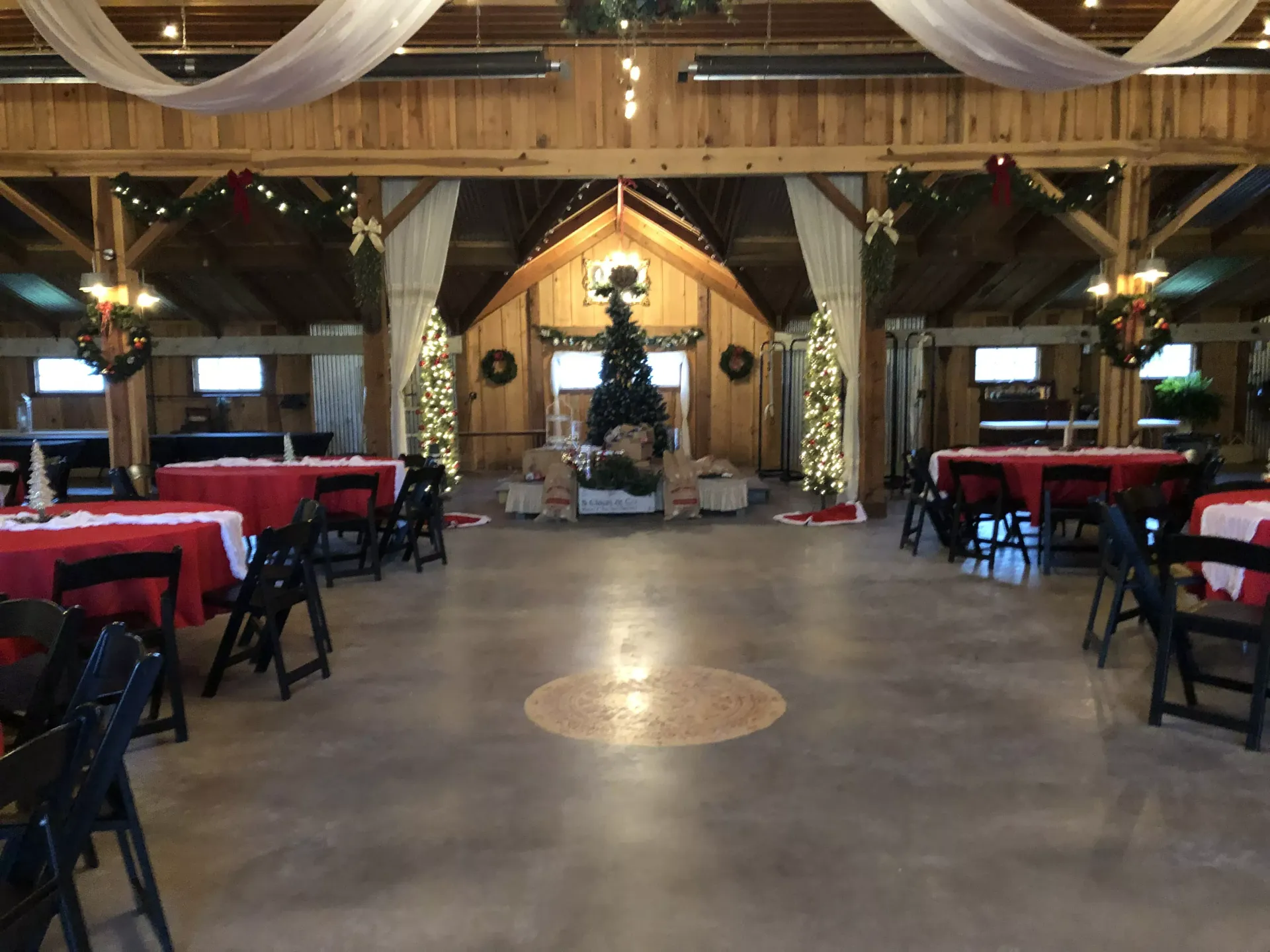 Rustic event hall with wooden beams, draped fabric, red tablecloths, and a central altar at the far end