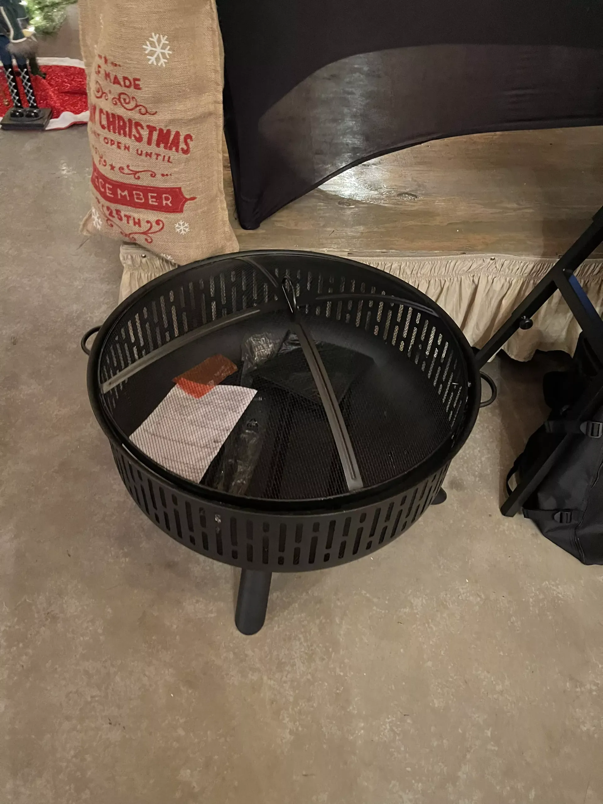 Black round metal fire pit or planter on a concrete floor indoors, beside a sack and furniture.