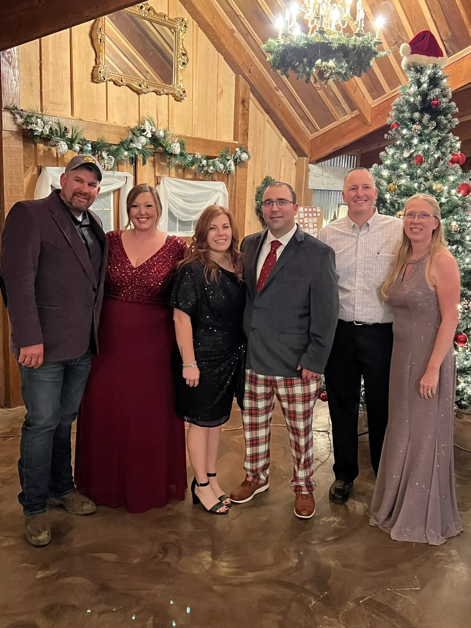 Six people posing together in a warmly lit wooden hall beside a decorated Christmas tree.