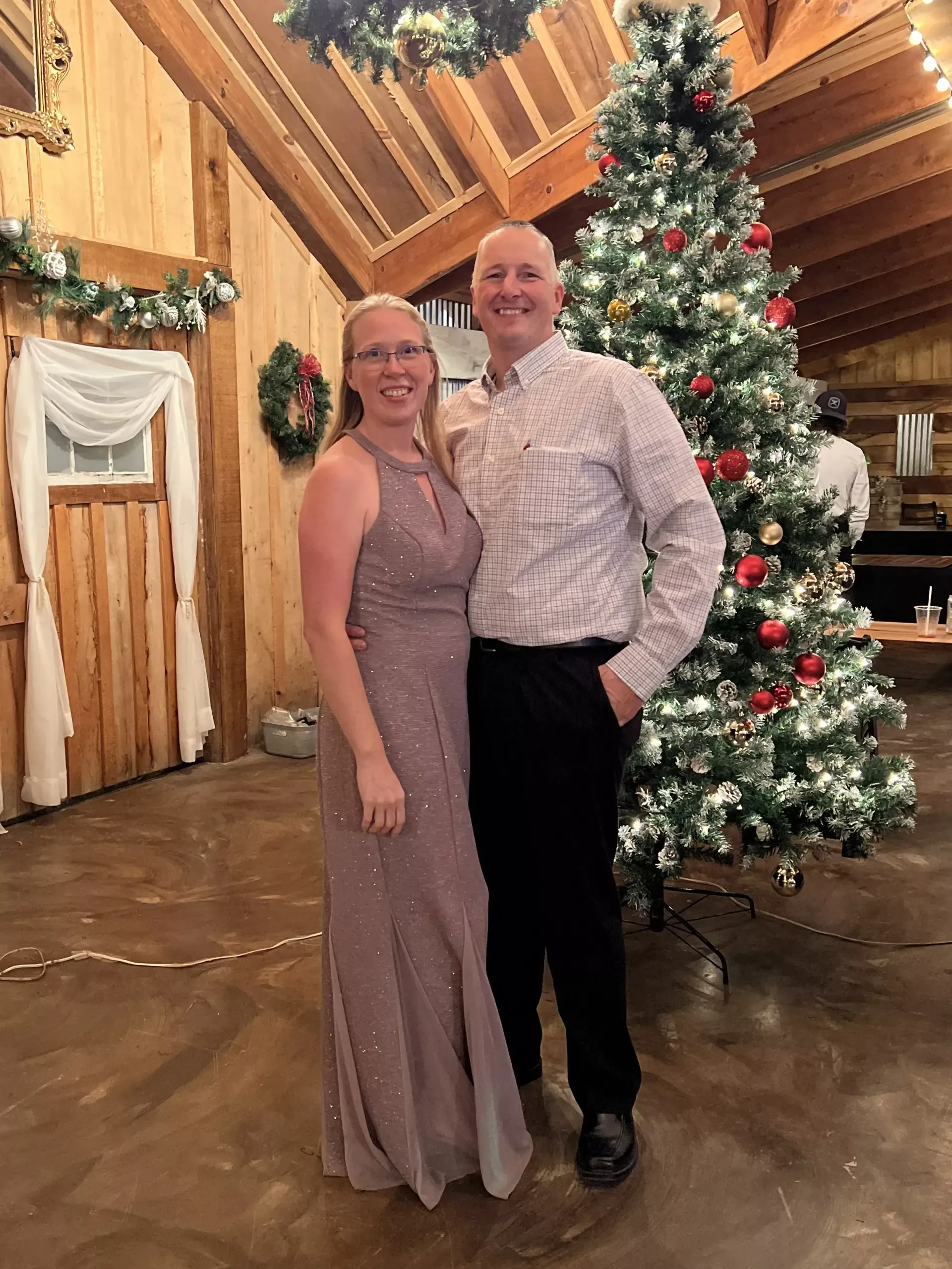 Two people posing together beside a decorated Christmas tree in a rustic hall.