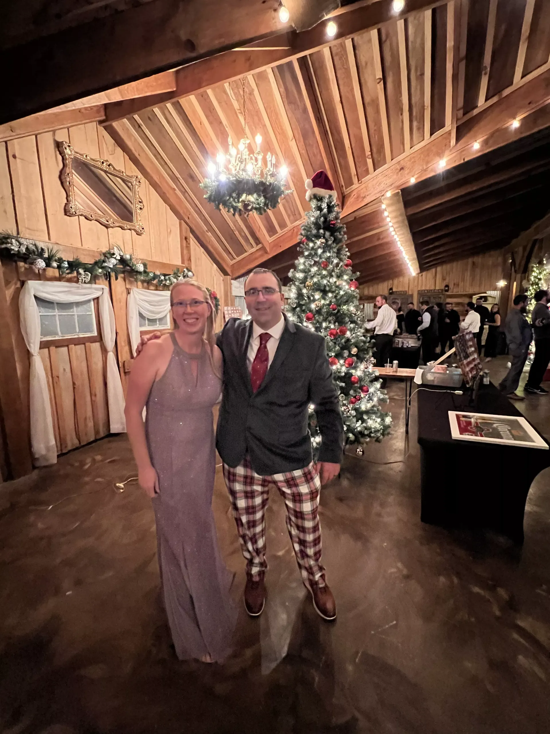 Two people posing indoors beside a decorated Christmas tree and chandeliers, with rustic wood beams and warm lights.