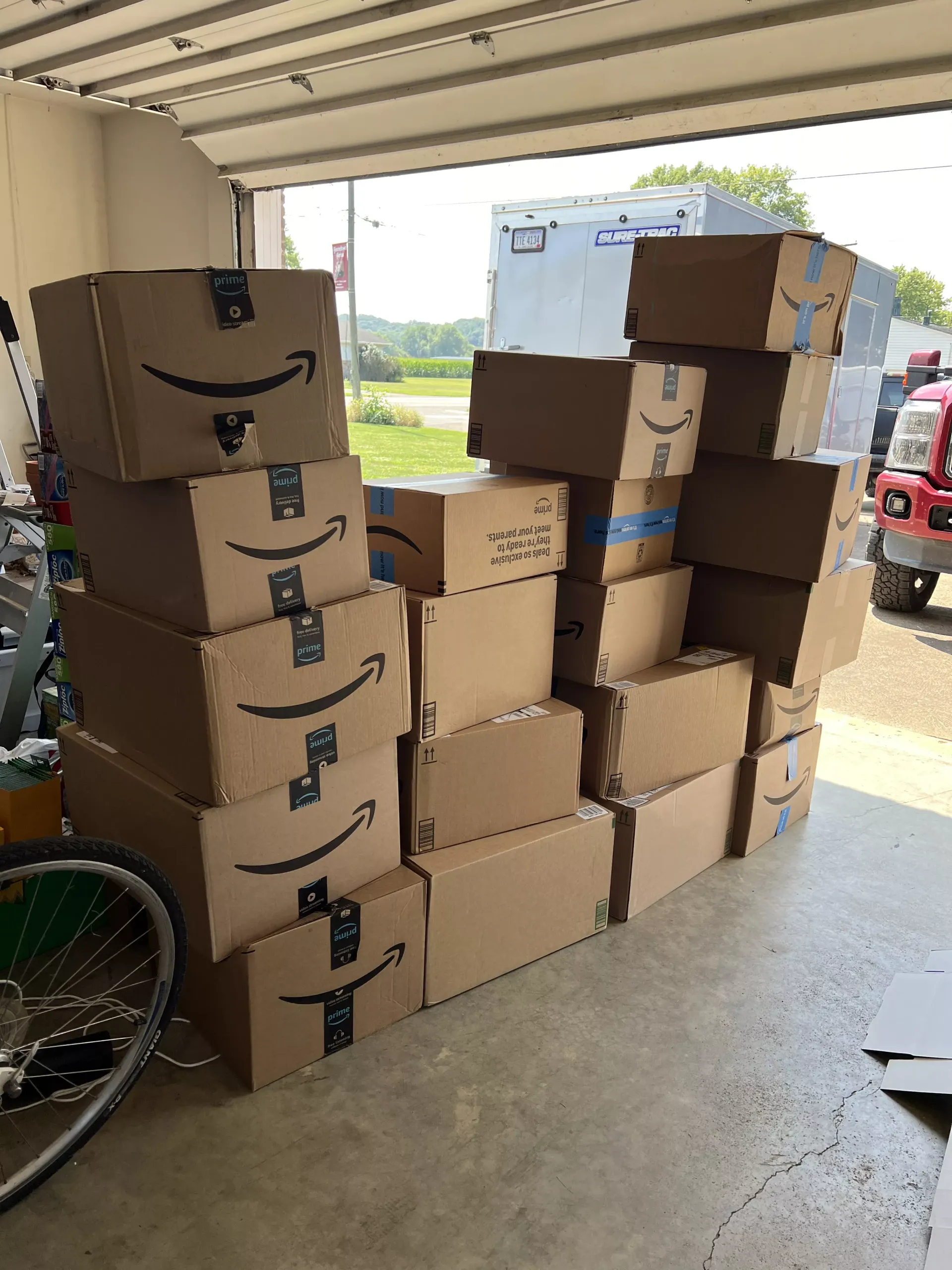 Stacks of Amazon boxes piled in a garage doorway, with a bicycle wheel visible on the left.