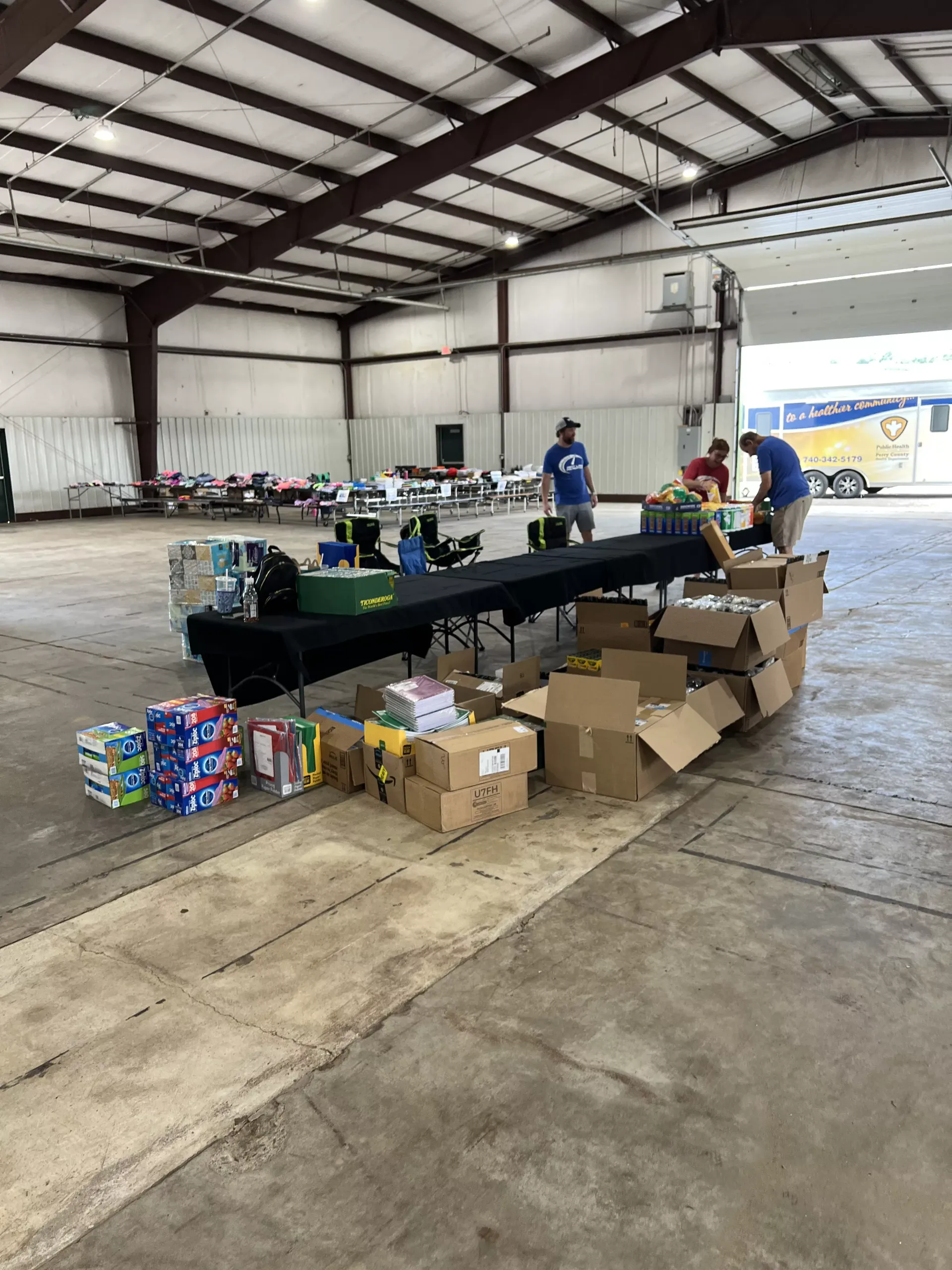 Warehouse volunteers sorting donated goods on long tables with boxes and supplies on the floor