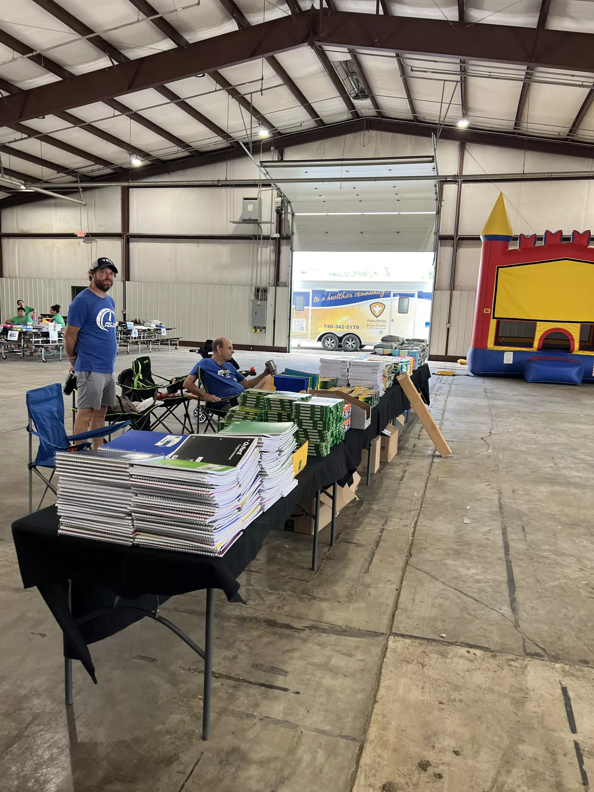 Indoor community event with tables of packaged goods and a projector screen in a large warehouse-like hall