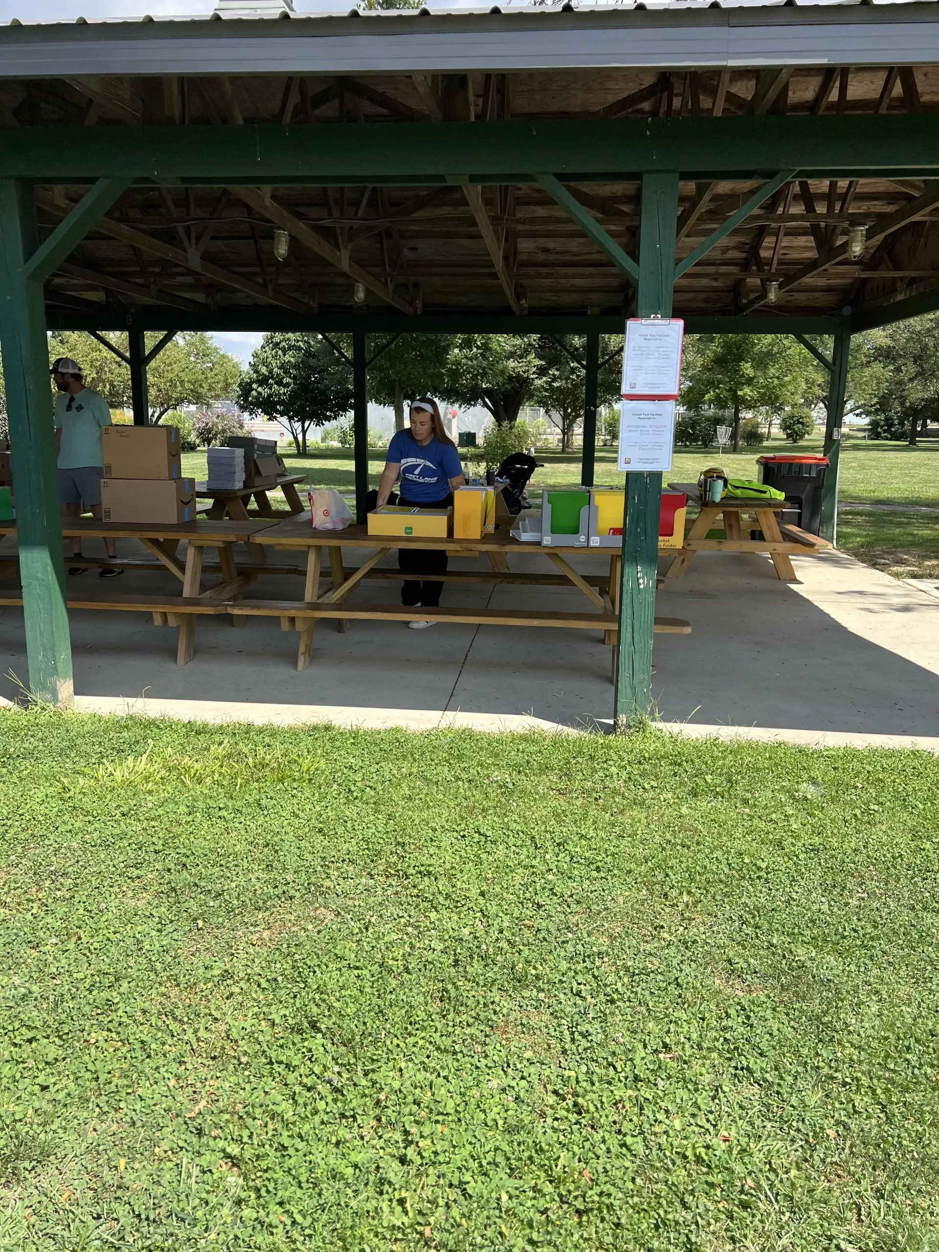 Covered picnic pavilion with people seated at tables in a grassy park area.