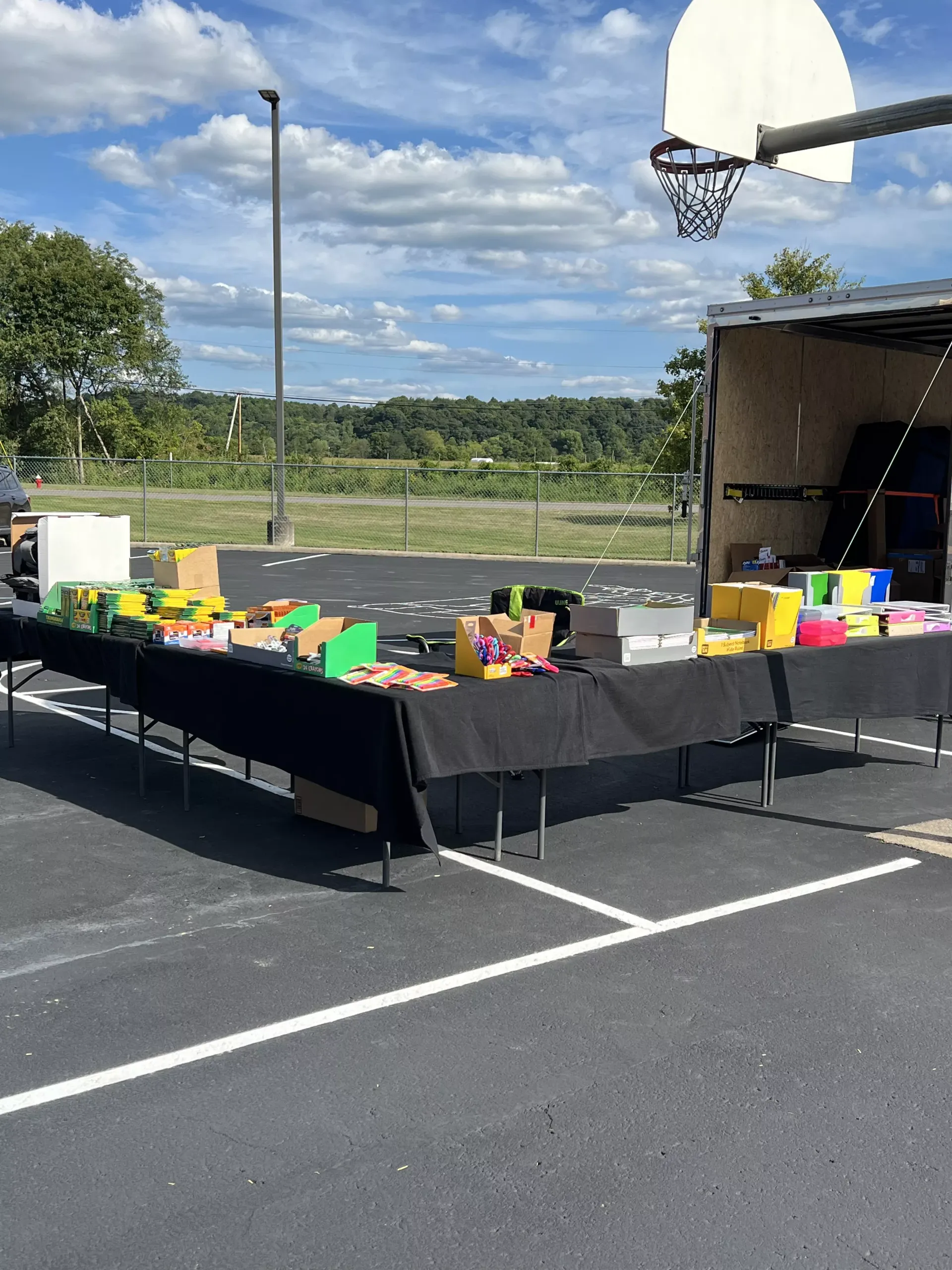 Outdoor basketball court with a long table of colorful boxes and items under a partly cloudy sky