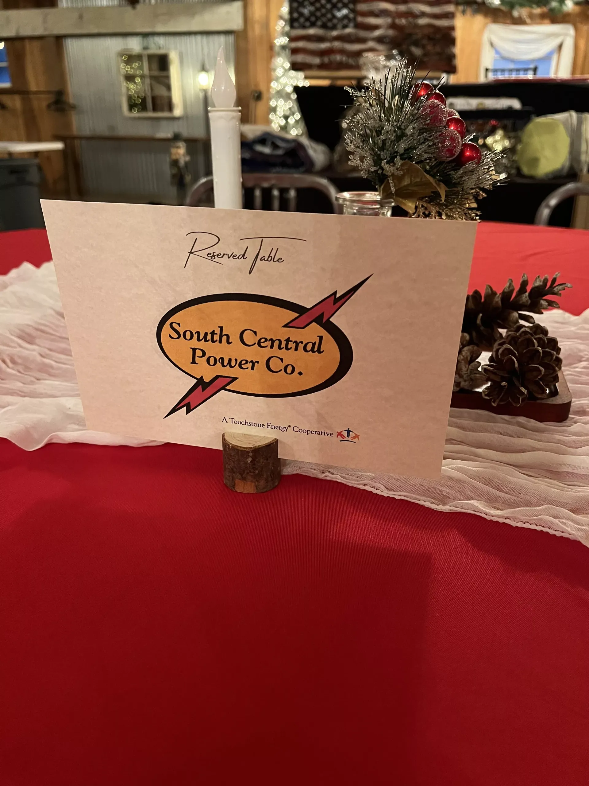 White card sign with logo on a red tablecloth, decorated with pinecones in an indoor event setting
