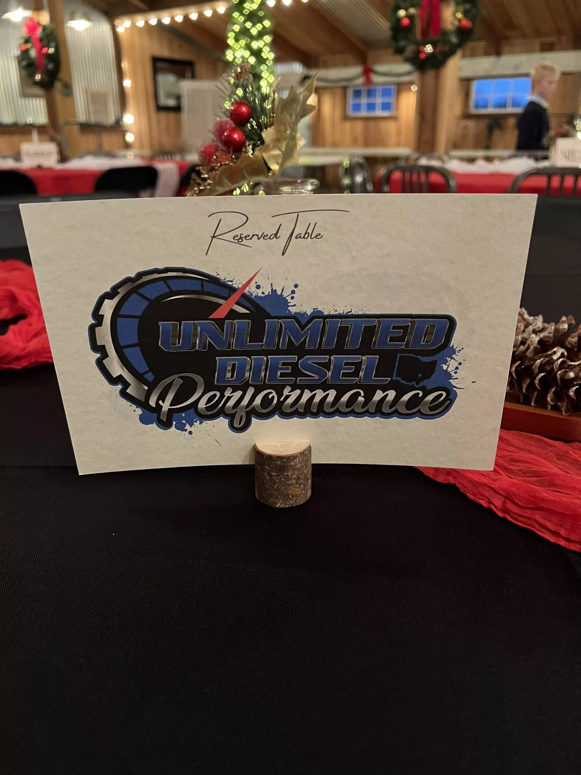 Table card with “Unlimited Diesel Performance” logo on a banquet table with red decor and blurred holiday lights.