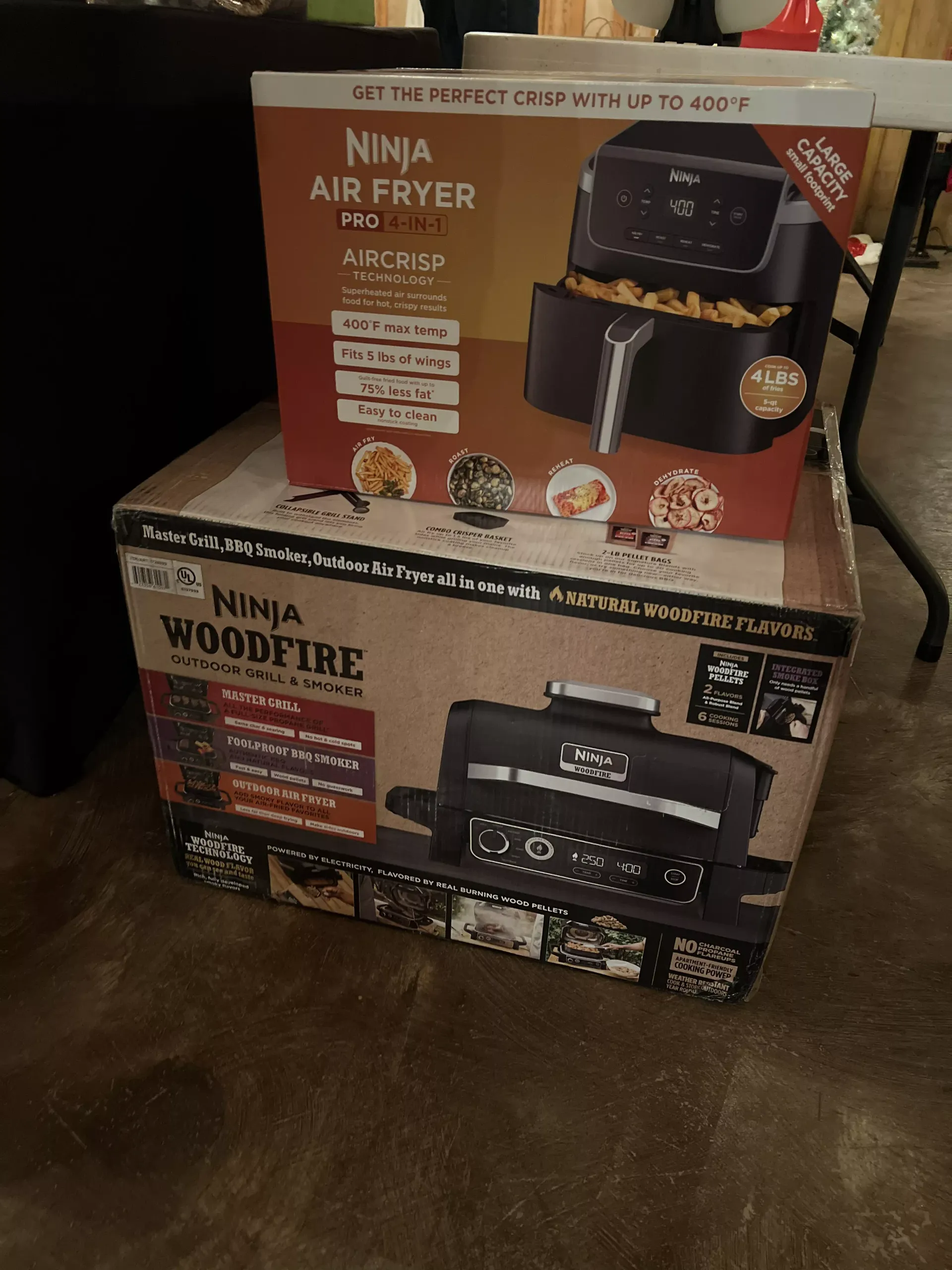 Two stacked Ninja appliance boxes on a wooden floor, including a black air fryer and a woodfire grill.