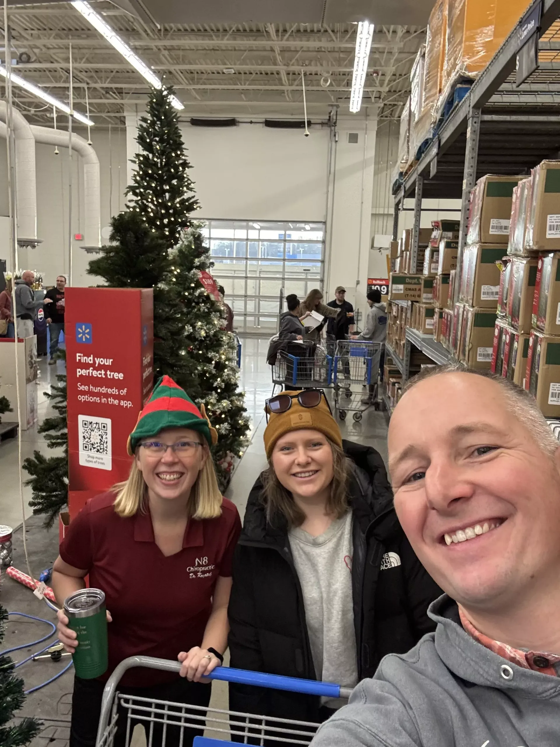 Three smiling shoppers in a warehouse store with a decorated Christmas tree behind them