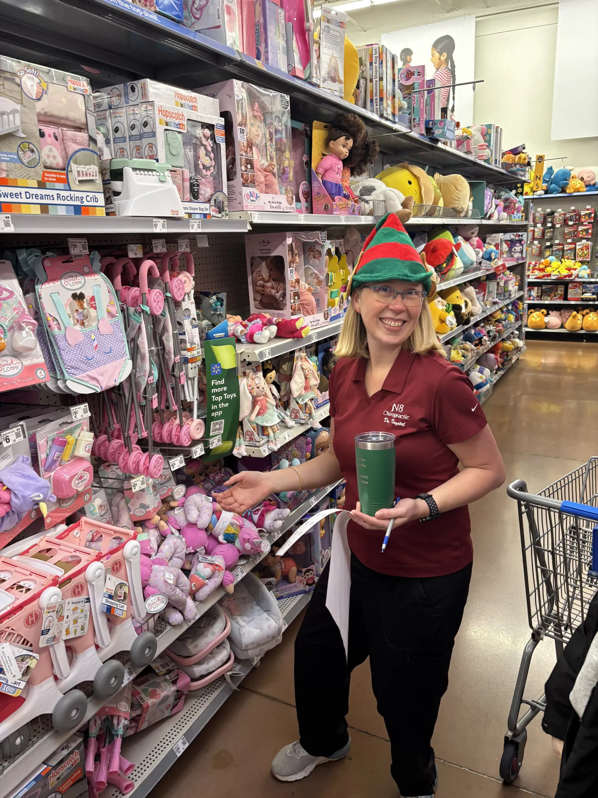 Woman in a toy store aisle holding a green tumbler and wearing a festive hat, surrounded by colorful toys.