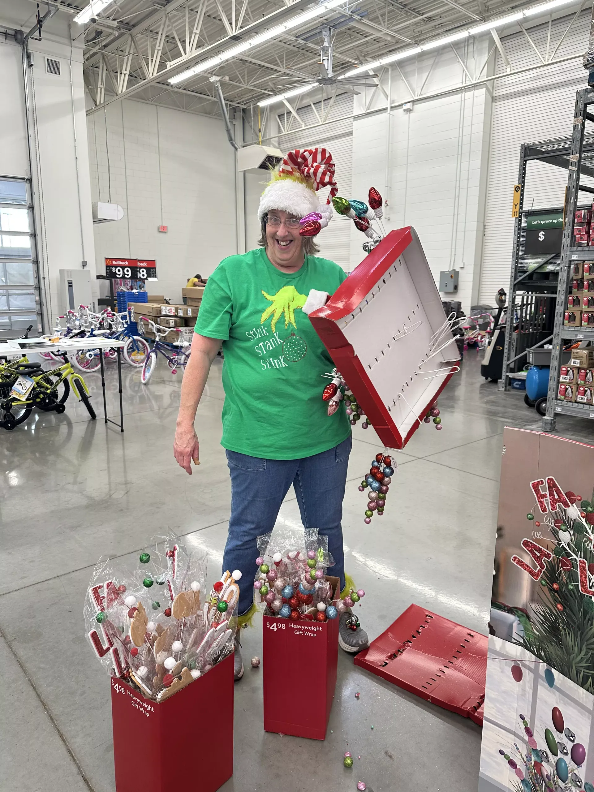 Person in green shirt and festive hat opening a red gift box in a warehouse-like space, surrounded by holiday decor