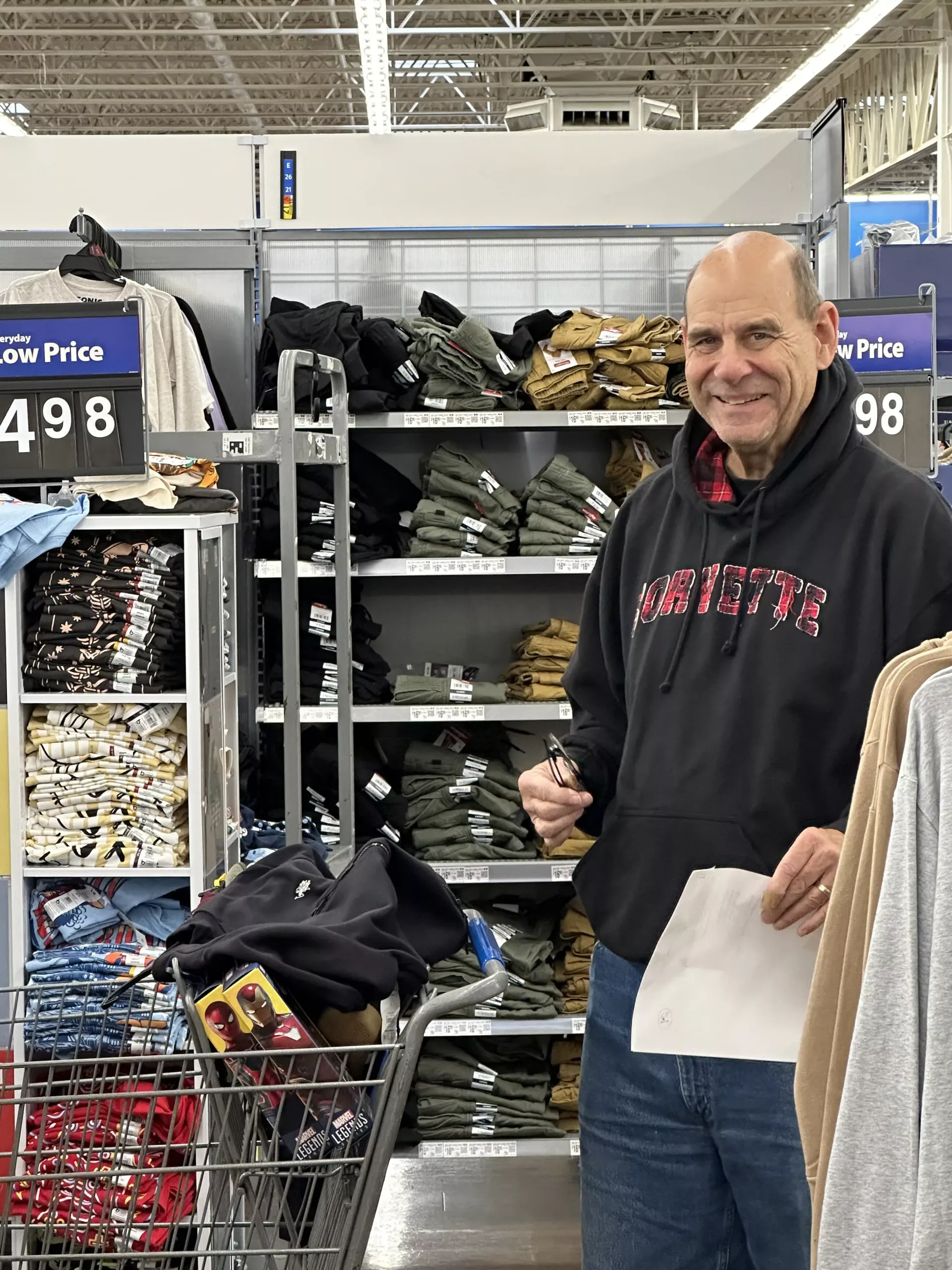 Man smiling in a store aisle beside a shopping cart and shelves of clothing, holding a paper.