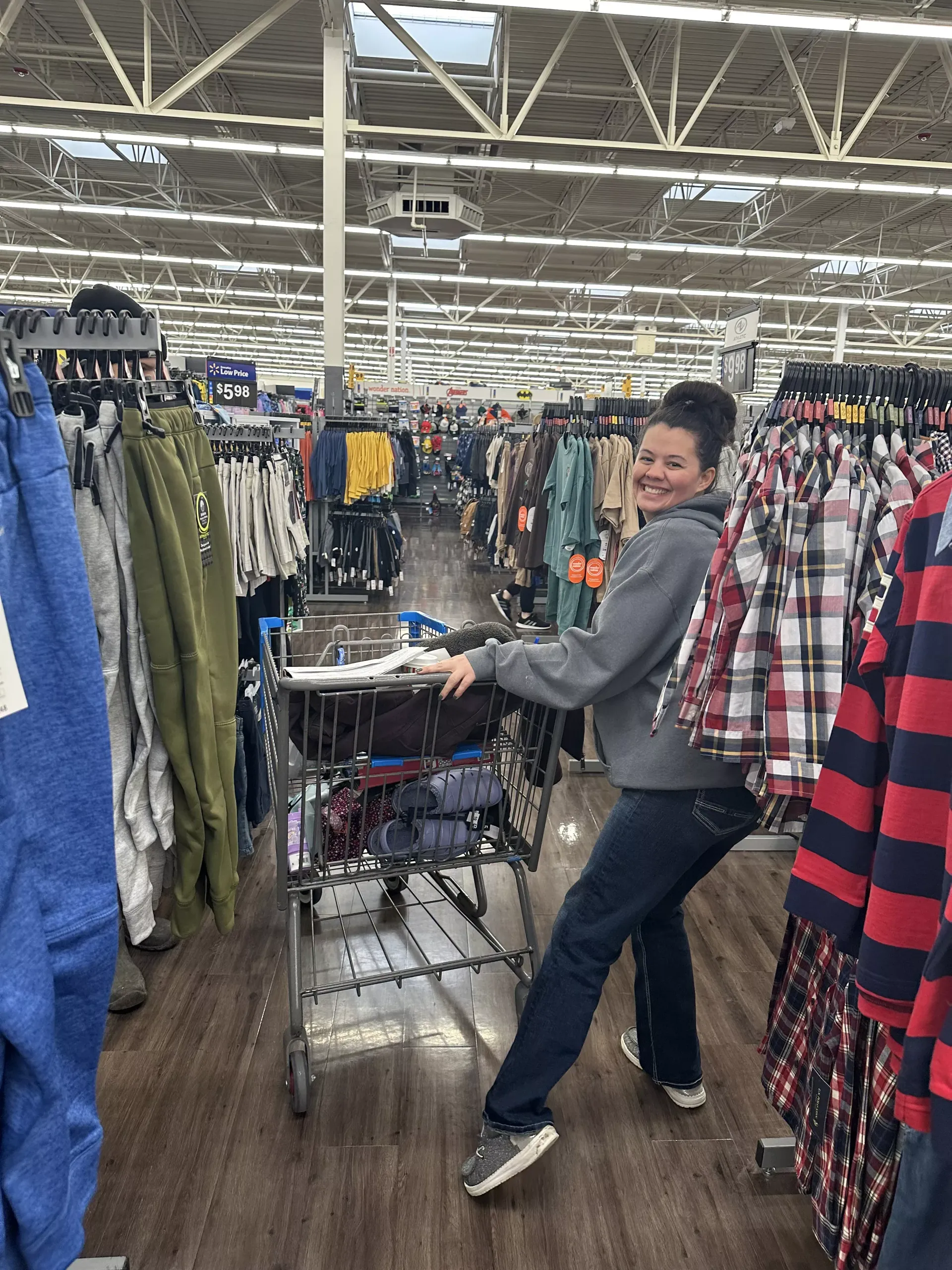 Person smiling in a clothing store aisle, pushing a cart between racks of shirts and jeans.