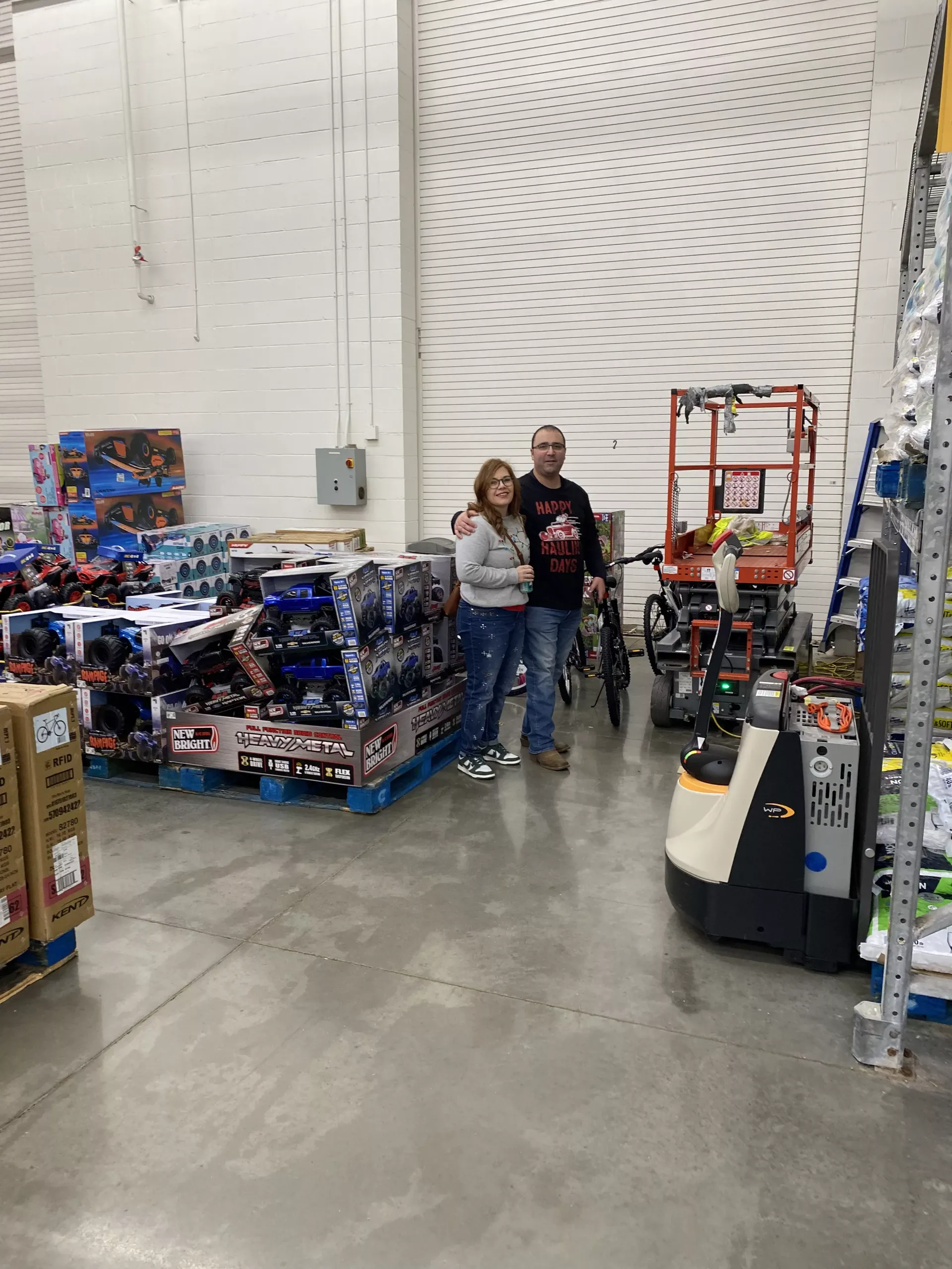Two people standing in a warehouse aisle beside stacked pallets of toy merchandise and carts