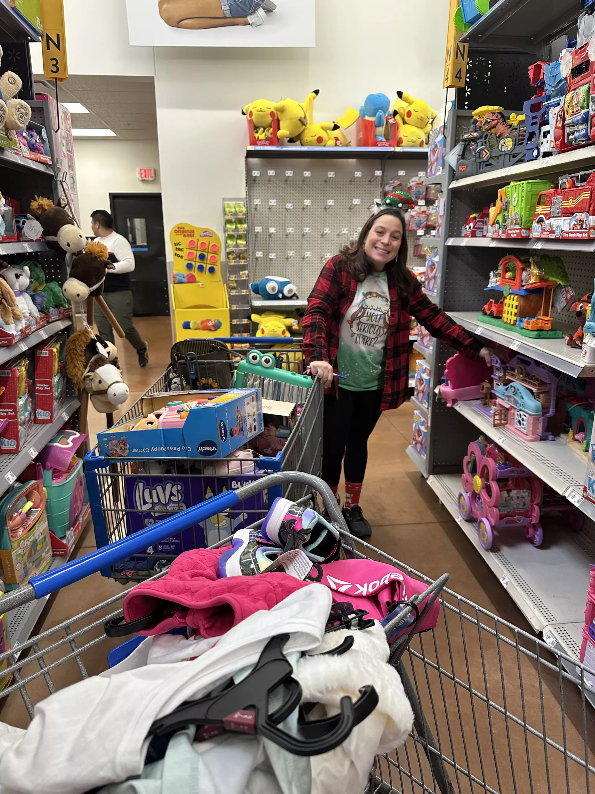 Woman smiling in a toy store aisle, leaning toward a shopping cart filled with items.