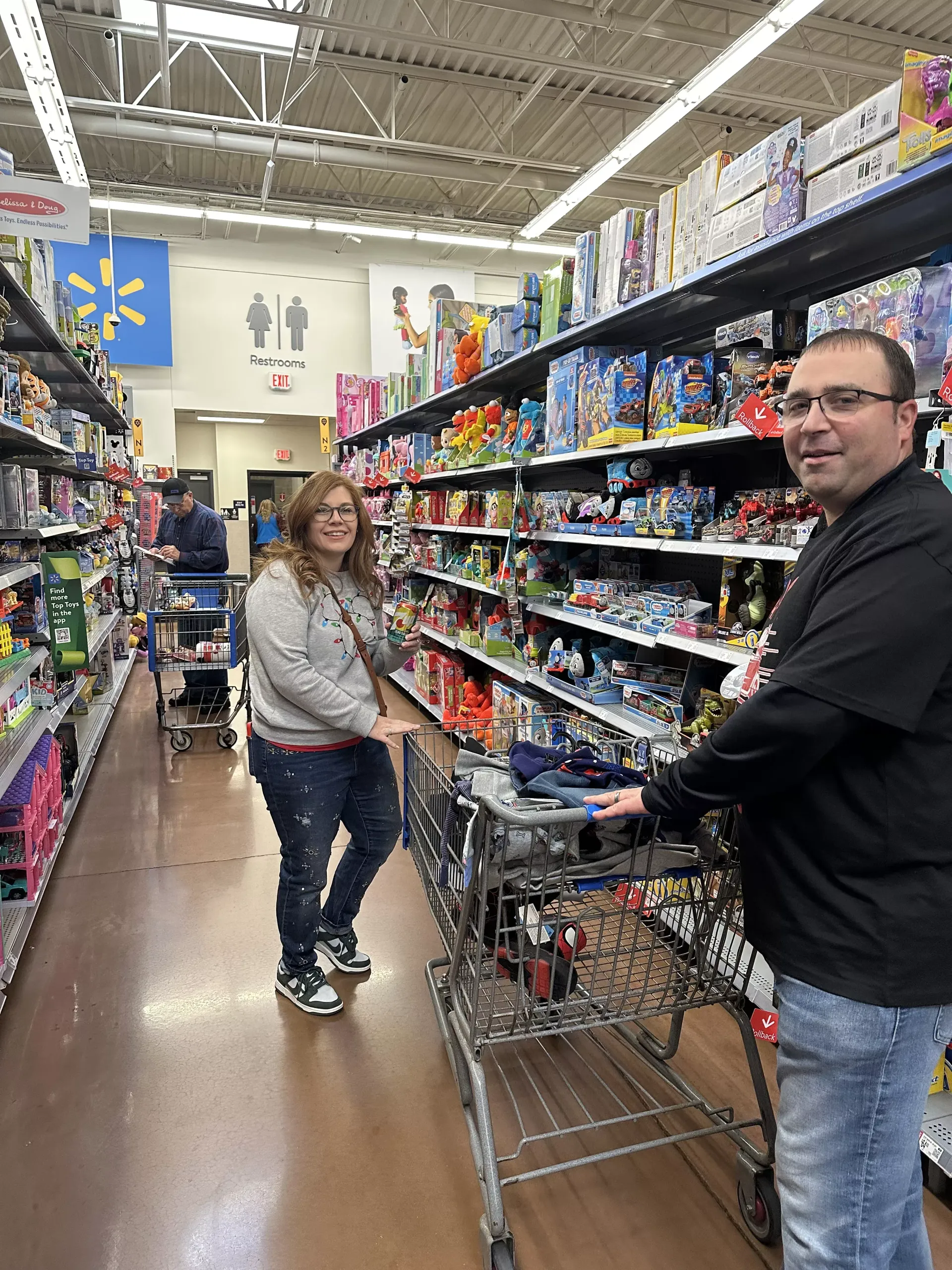 Two people with shopping carts in a toy store aisle, one smiling at the camera.