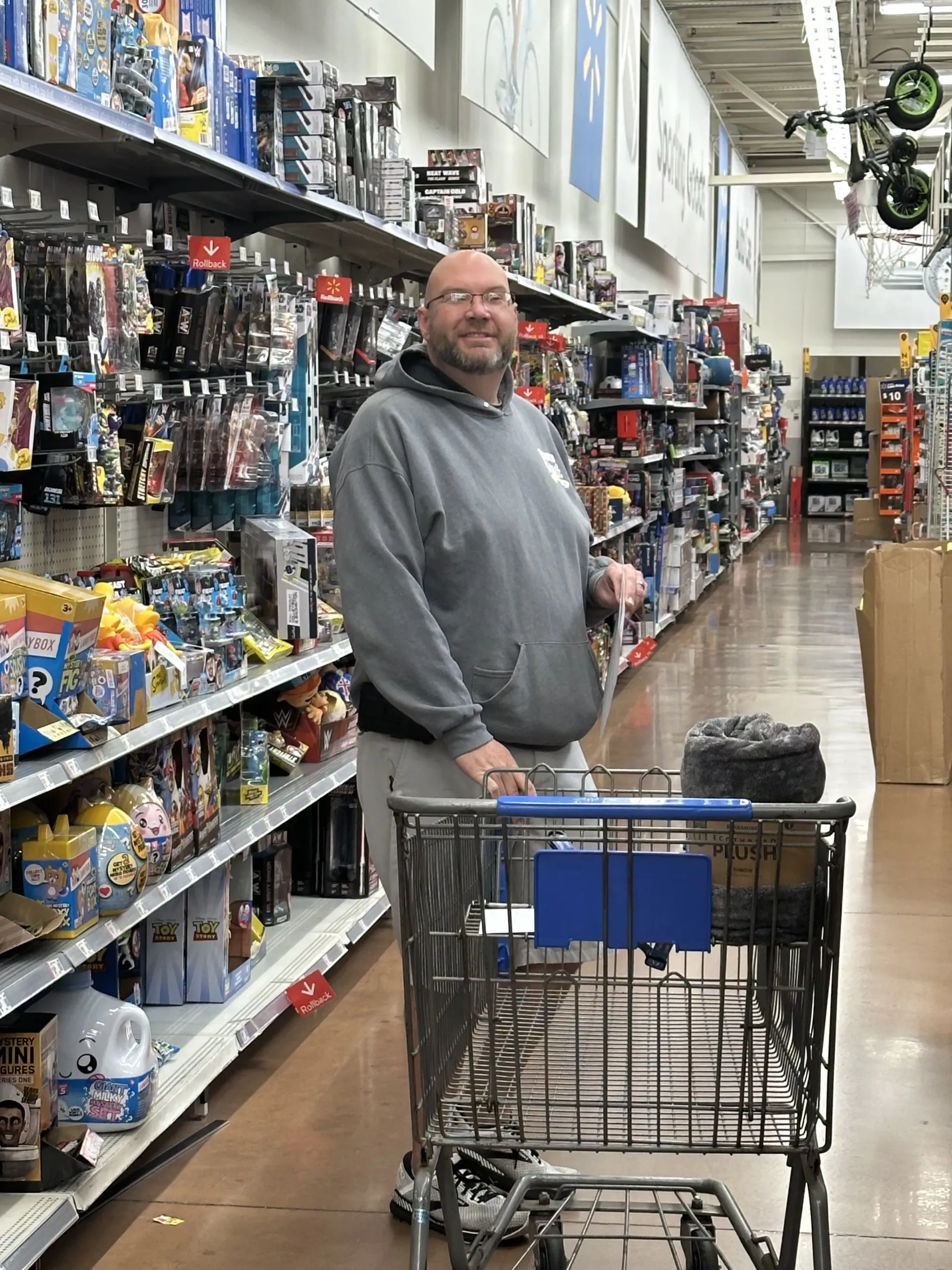 Man standing with a shopping cart in a grocery store aisle, wearing a gray hoodie.