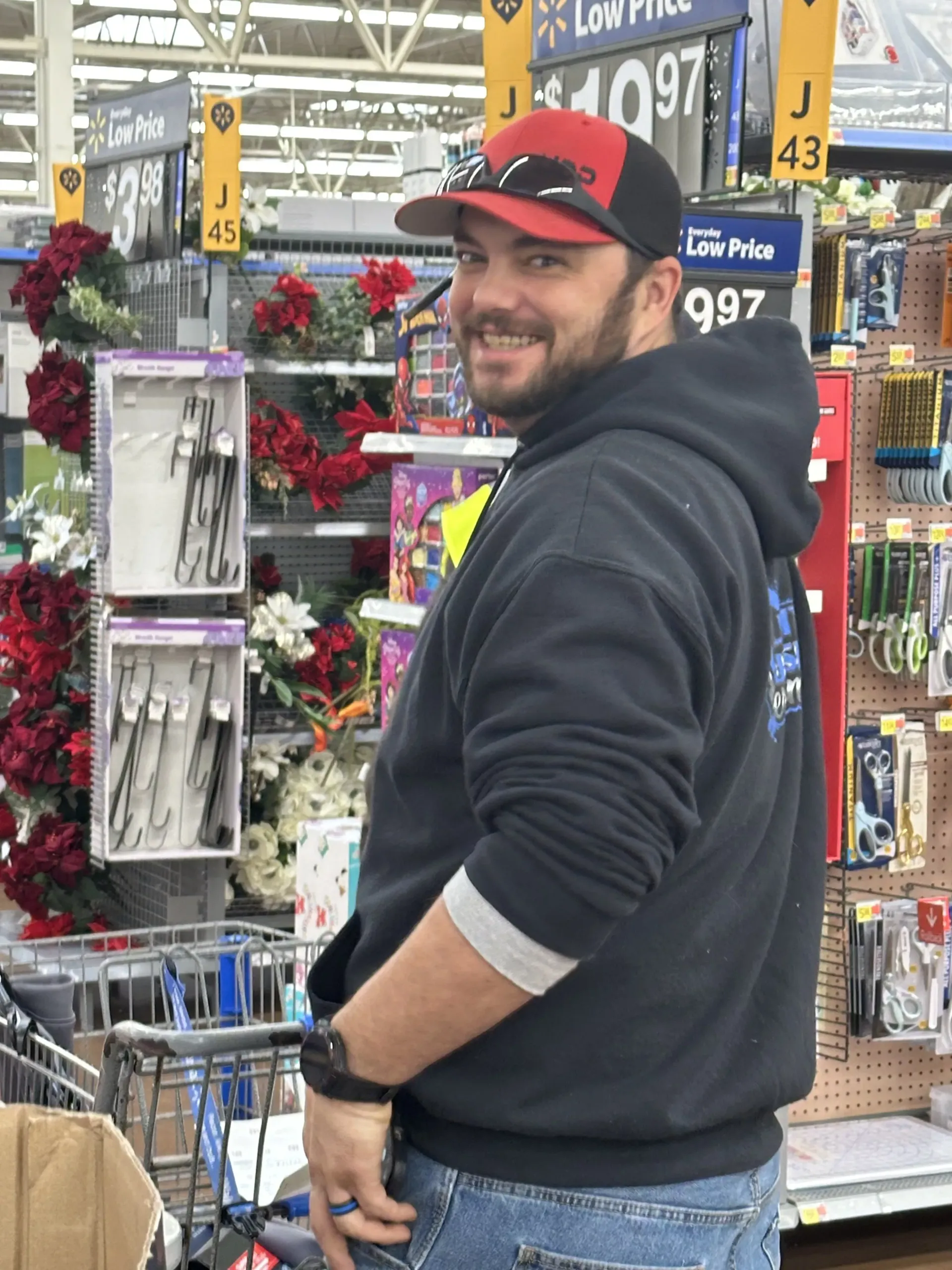 Smiling man in black hoodie and red cap standing in a store aisle beside shopping carts and holiday decorations