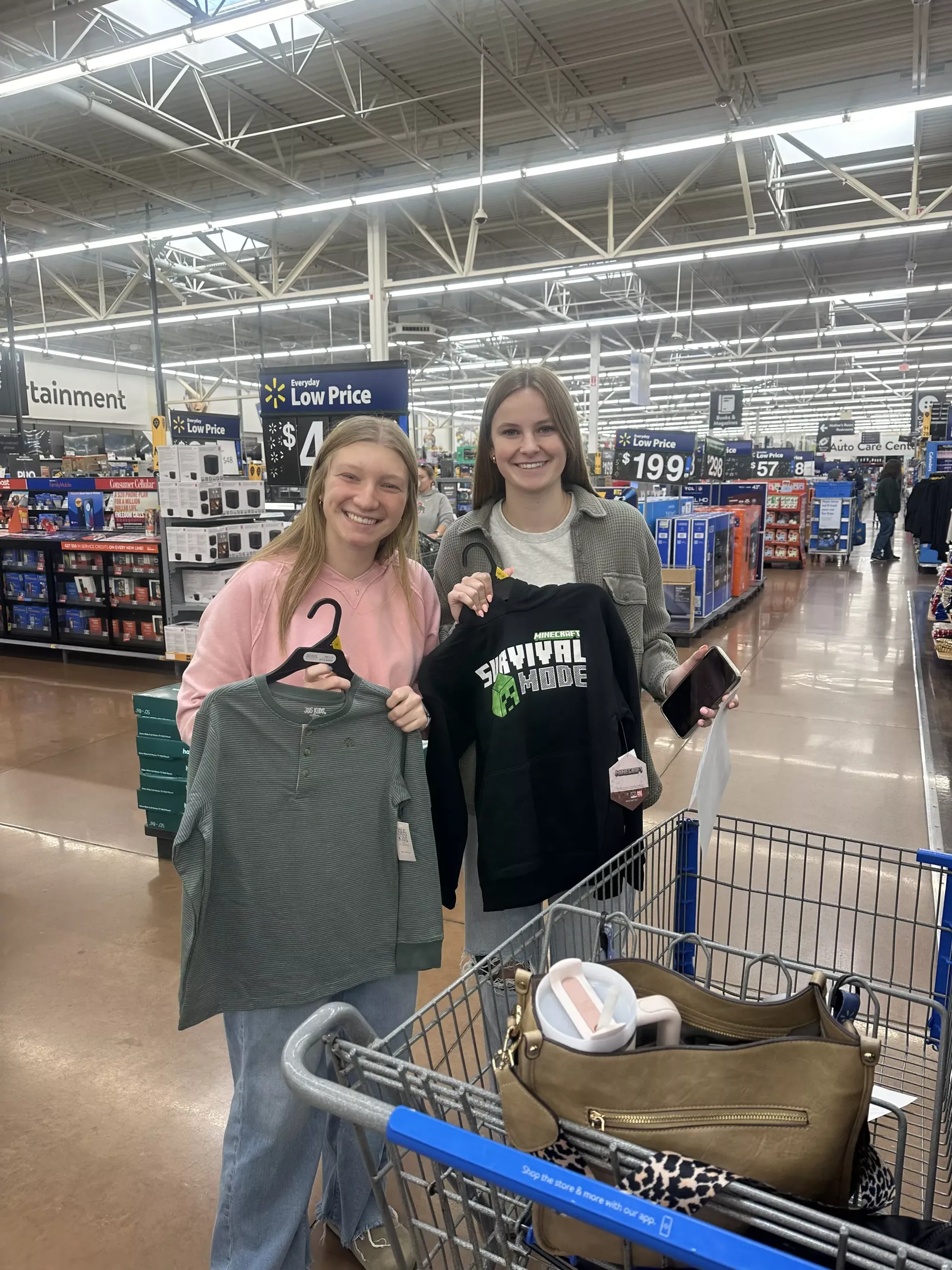 Two shoppers holding shirts in a brightly lit store aisle beside a shopping cart.