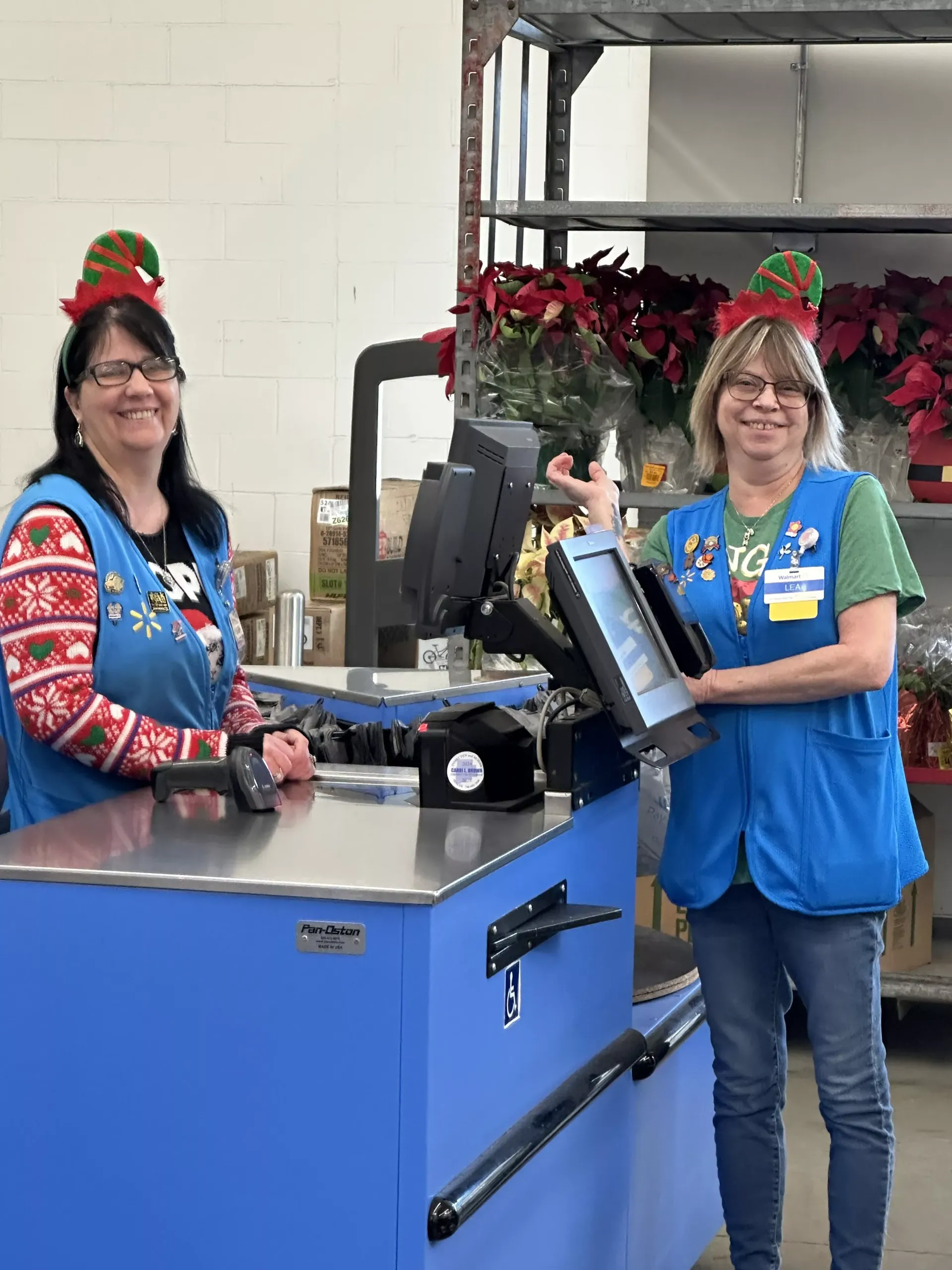 Two smiling workers in blue aprons at a checkout counter in a store, with holiday hats and poinsettias nearby