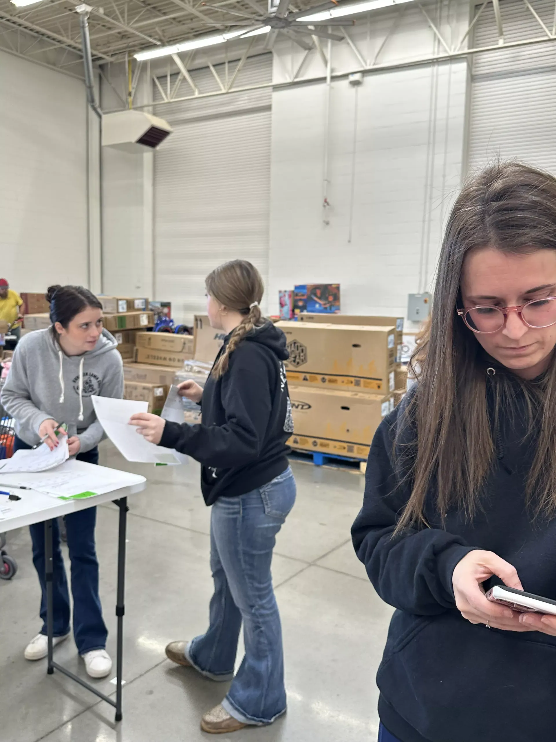 People sorting papers and using a phone in a warehouse with stacked boxes and tables