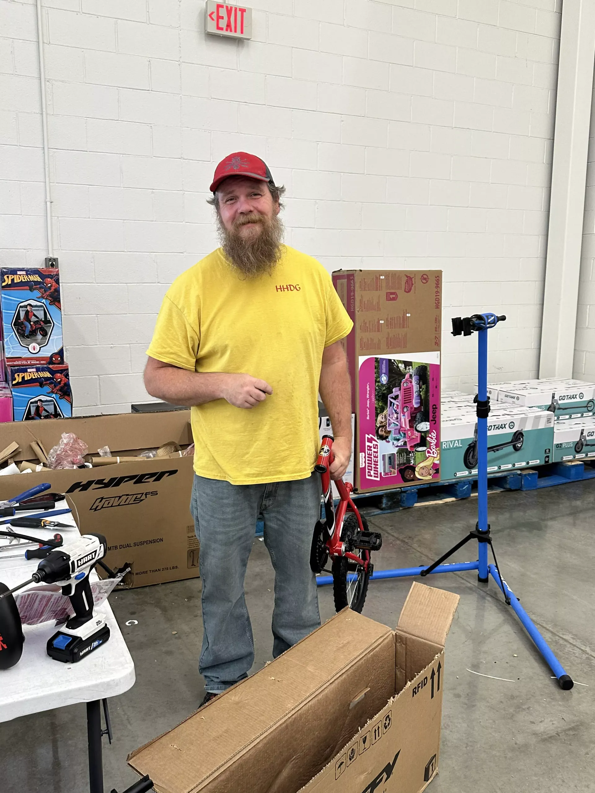 Man in a yellow shirt and red cap standing by boxes and event gear in a warehouse-like room