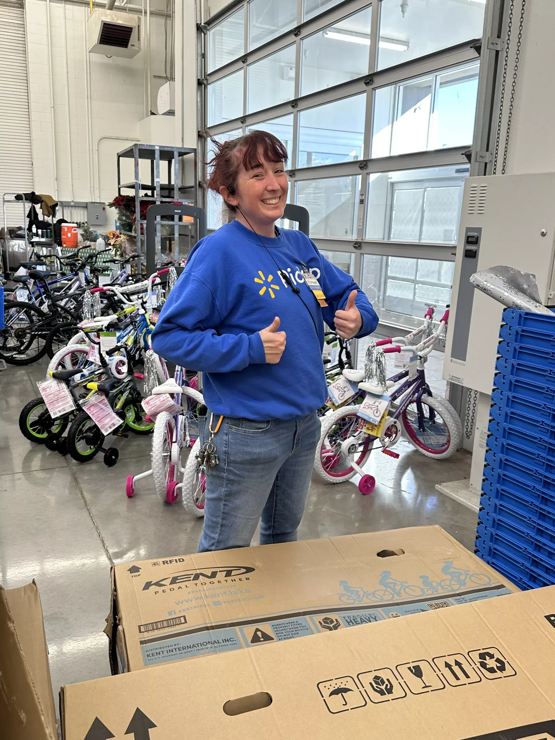 Smiling person in a blue hoodie giving thumbs up beside stacked bicycle boxes in a bright shop