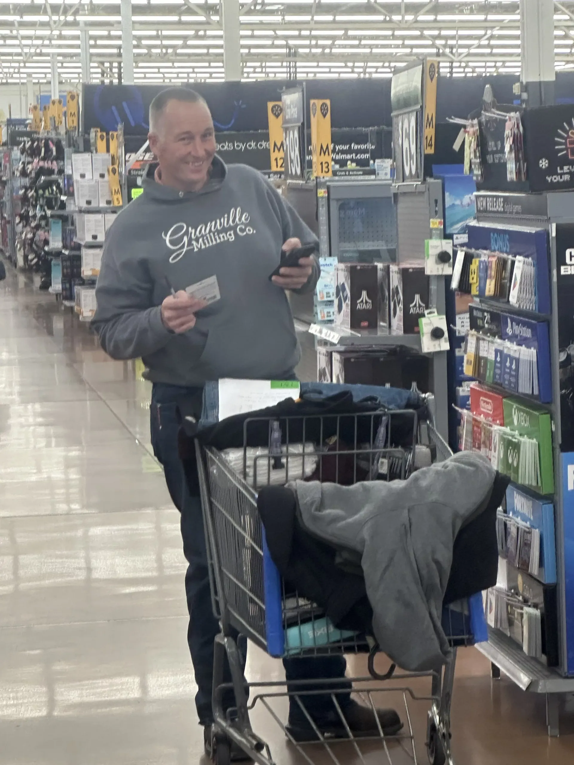 Man in a store aisle pushing a shopping cart filled with clothes and holding a folded item
