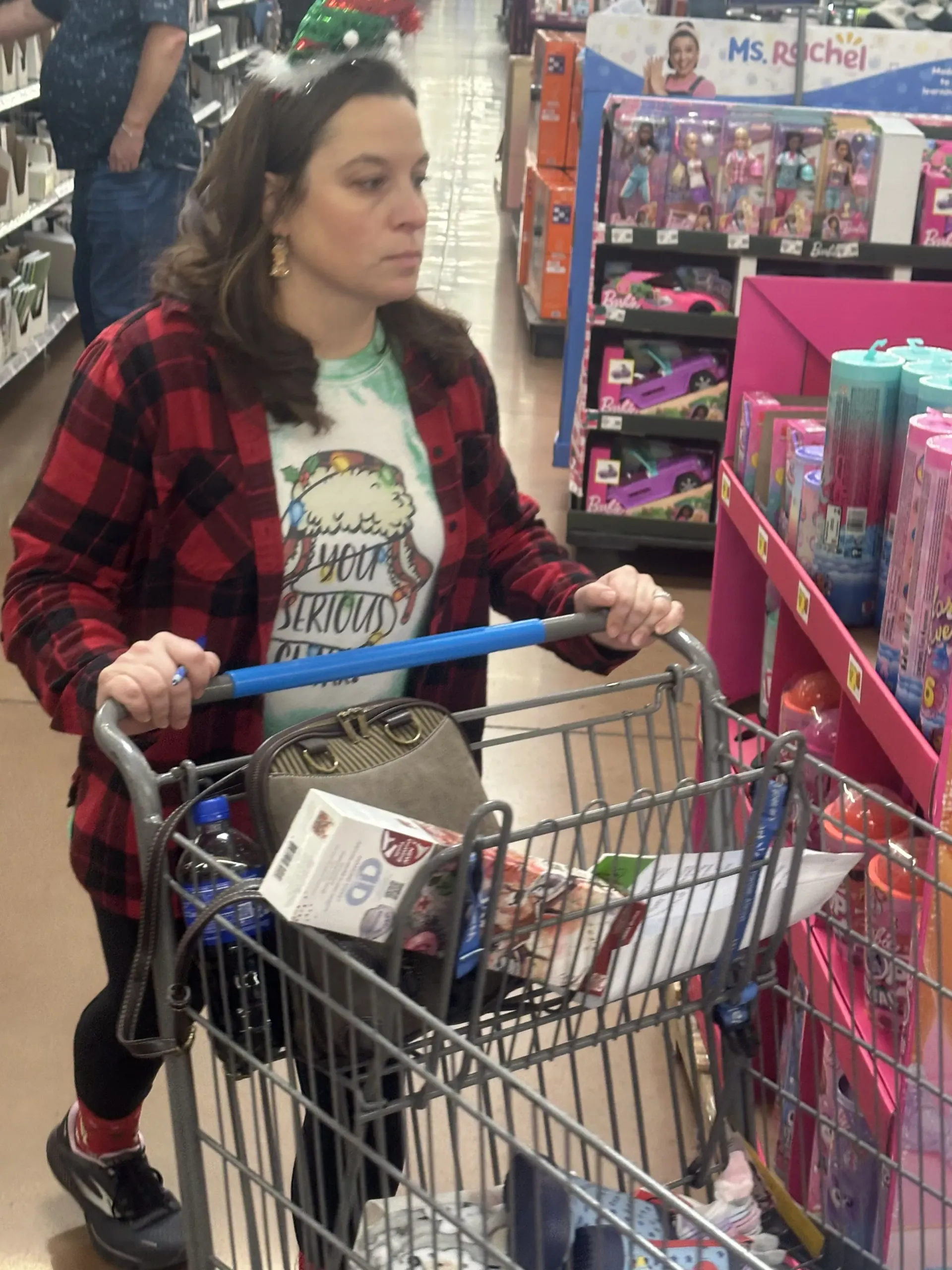 Woman in red plaid jacket pushing a shopping cart in a toy store aisle.