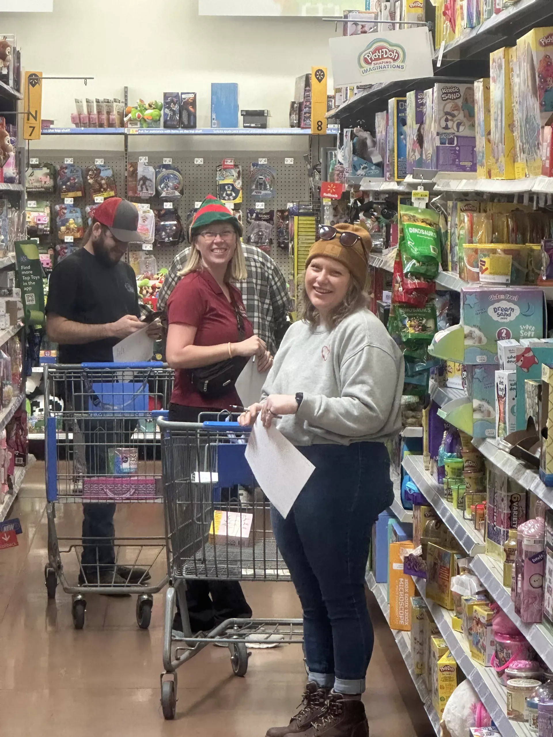Three shoppers with carts in a colorful toy store aisle, one smiling at the camera.