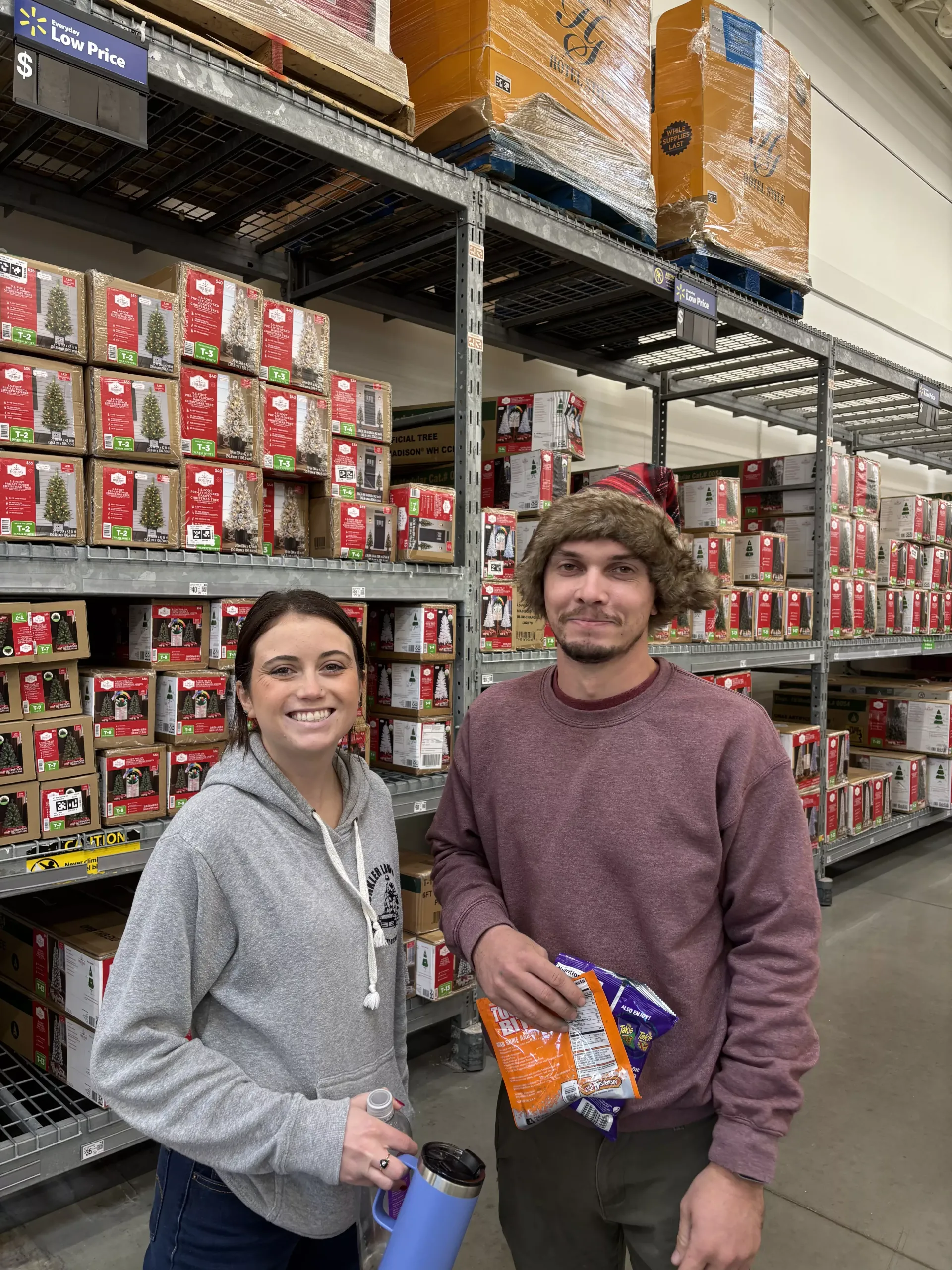Two people smiling in a warehouse aisle holding snacks, with stacked boxes on shelves behind them