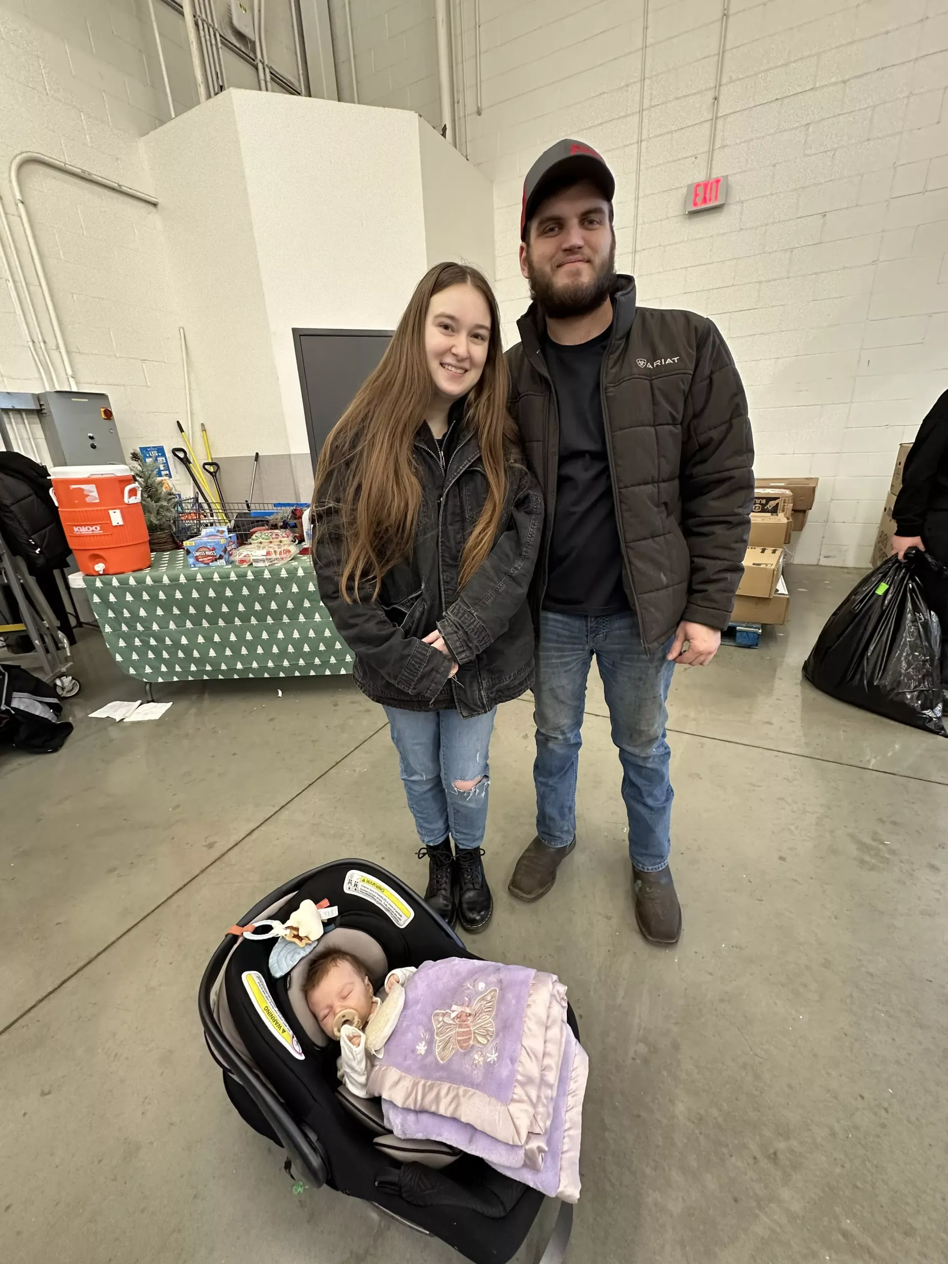 Couple standing indoors beside a sleeping baby in a car seat, with bags and tables in the background.