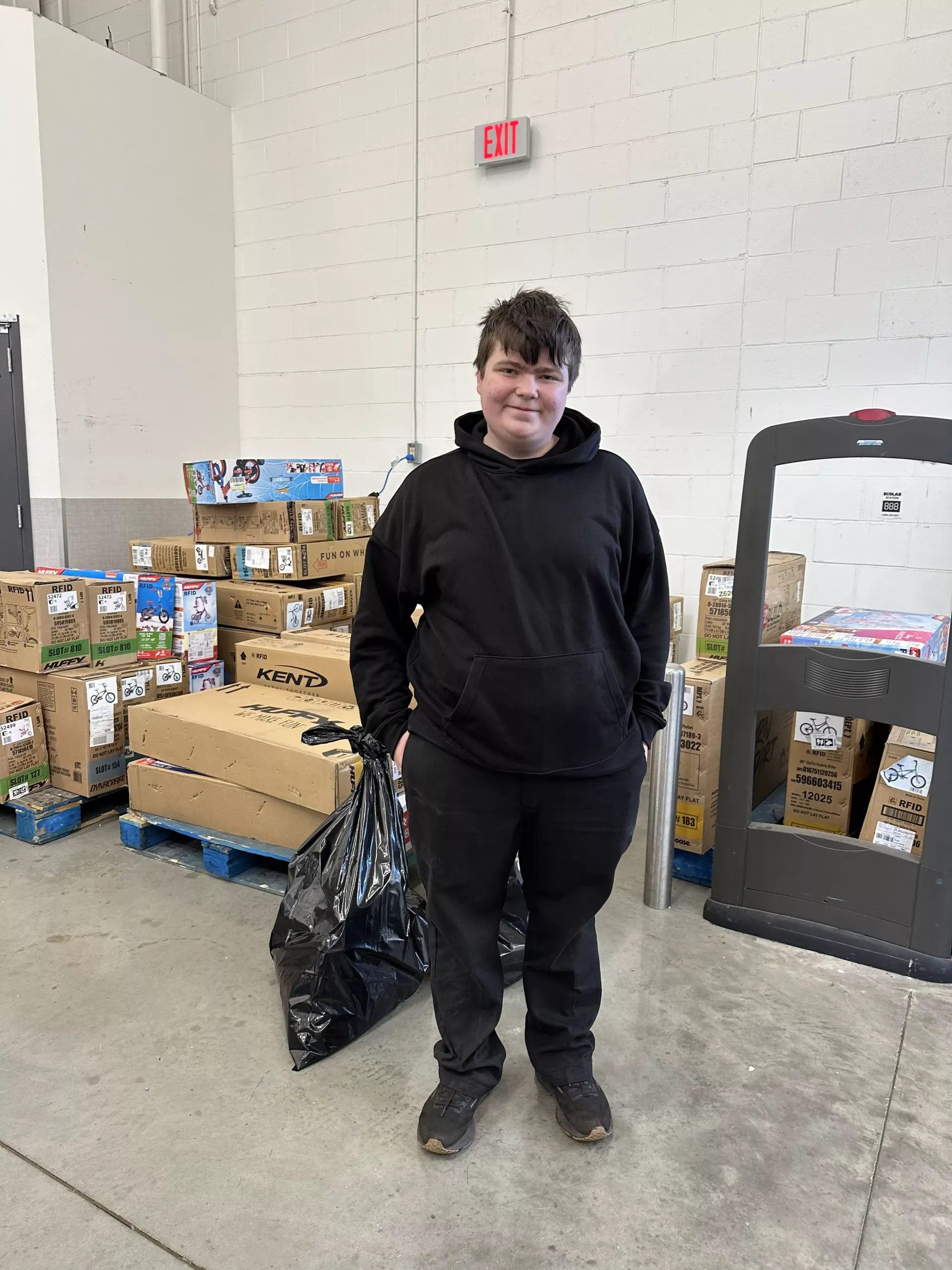Person in a black hoodie standing in a warehouse beside stacked boxes and a pallet jack.