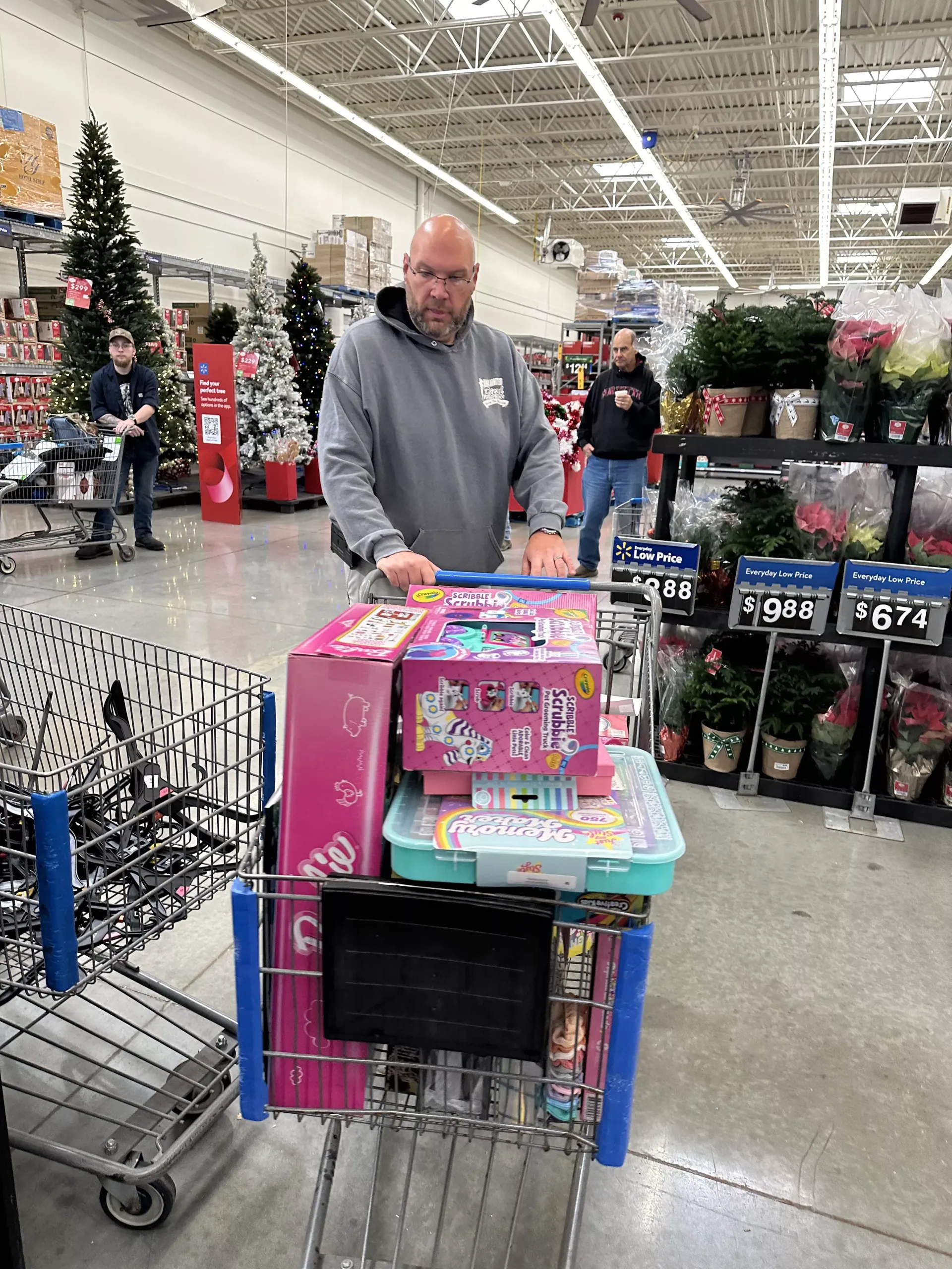 Man in a store aisle pushing a shopping cart loaded with a pink toy box and other items