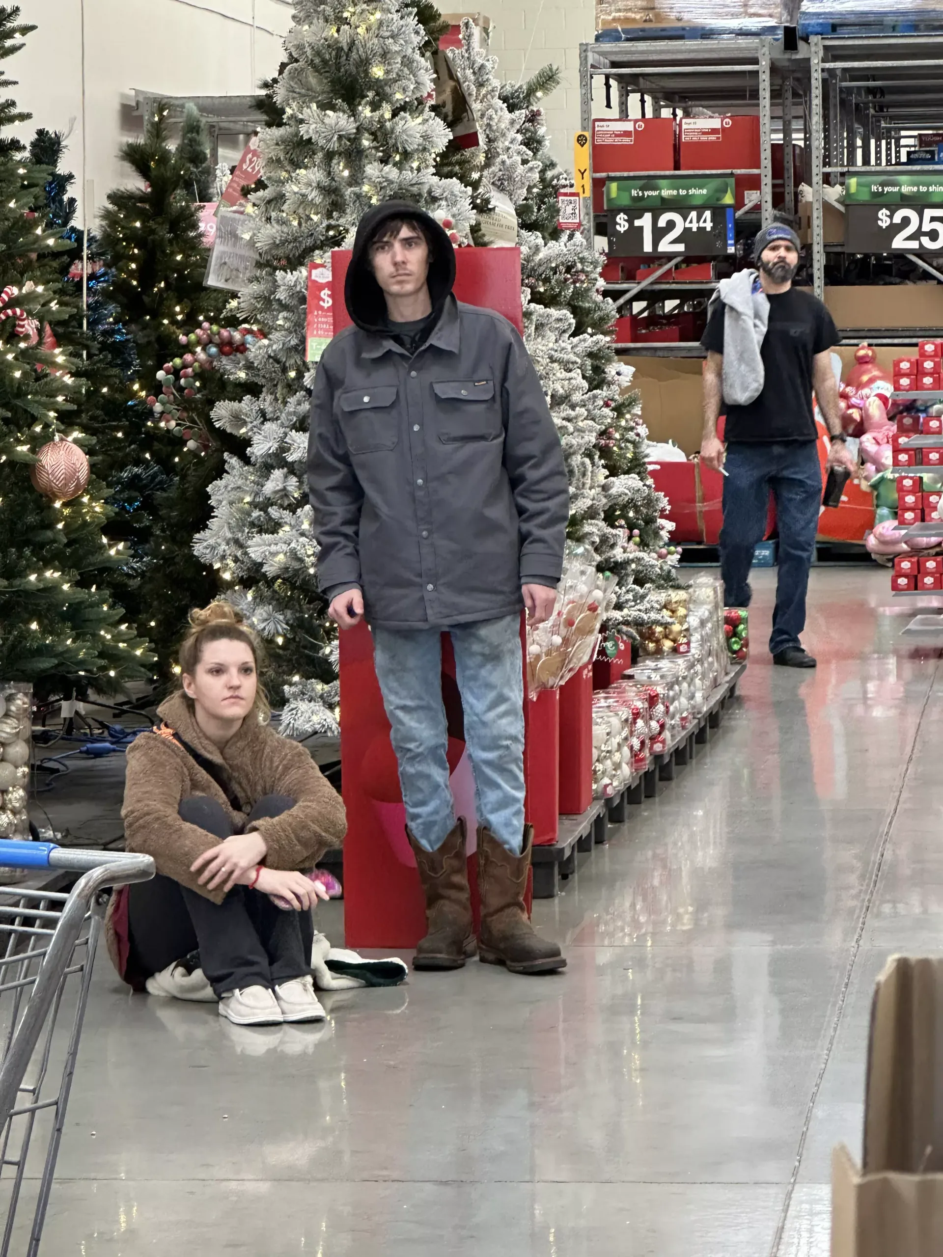 Two people by a decorated Christmas tree in a store aisle, with holiday decorations and price signs nearby