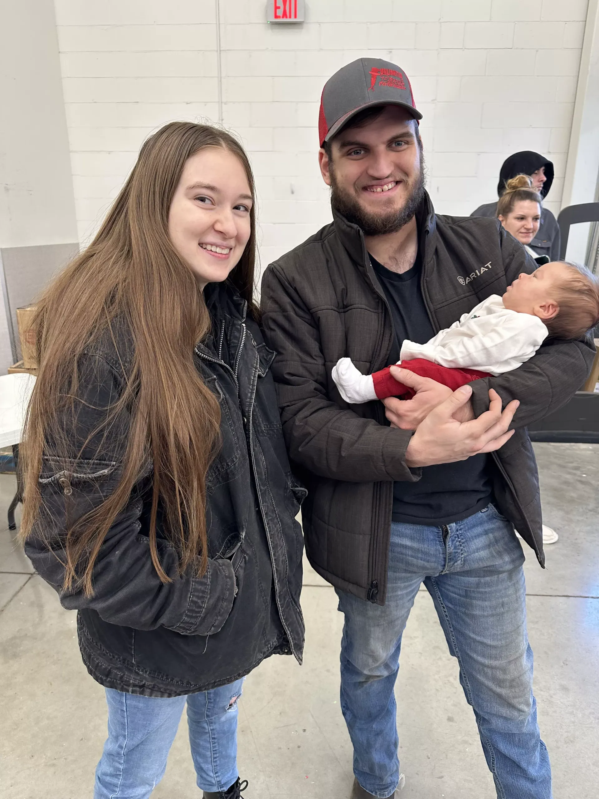 Two people smiling and holding a baby in a gymnasium or school hallway