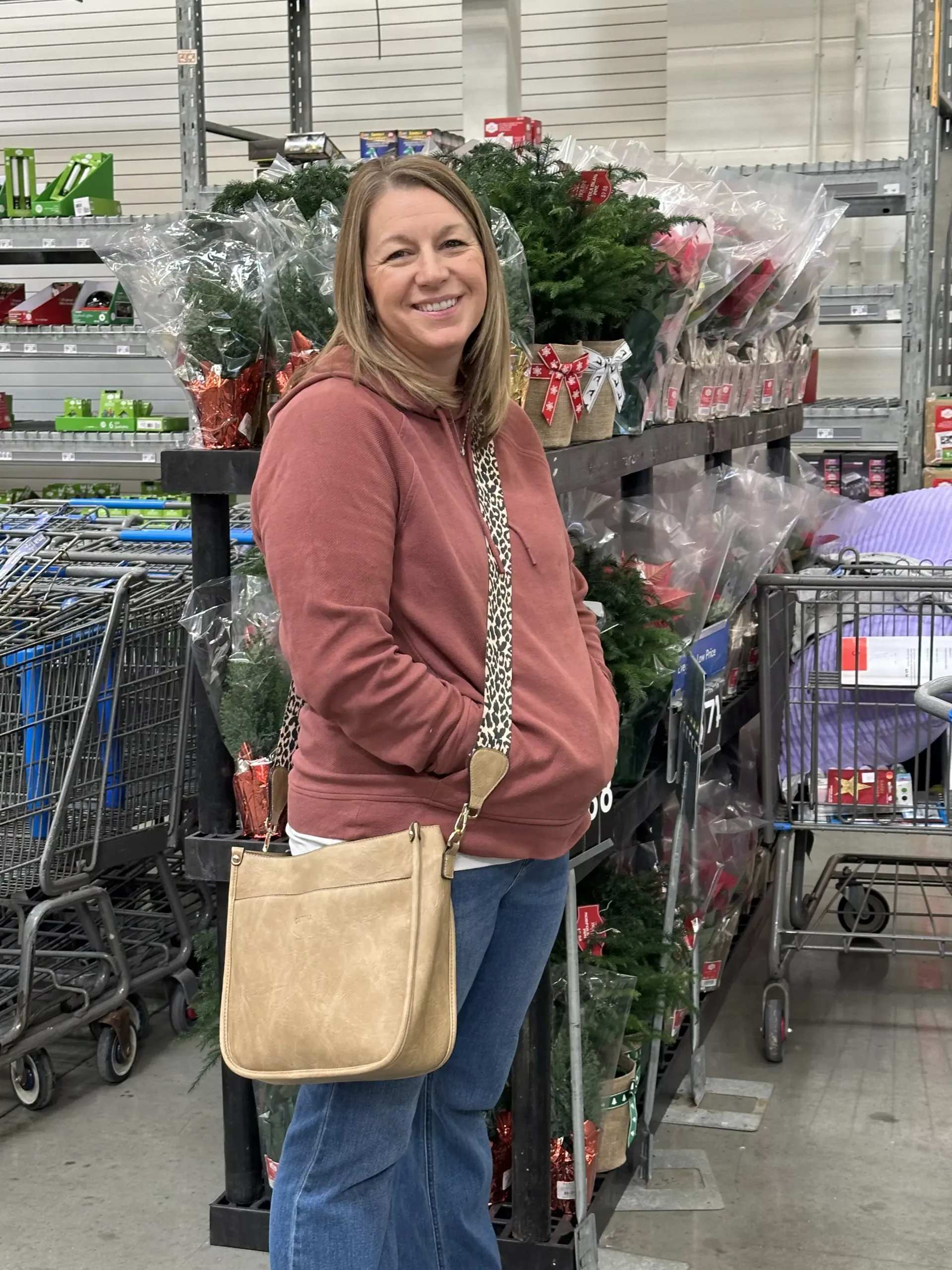 Woman smiling in a store aisle beside flower displays, wearing a pink sweater and beige crossbody bag.