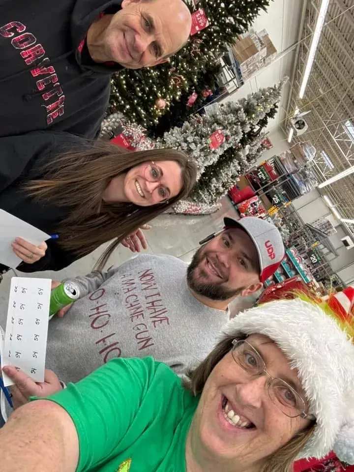 Four smiling people pose in a store’s holiday aisle with decorated Christmas trees.
