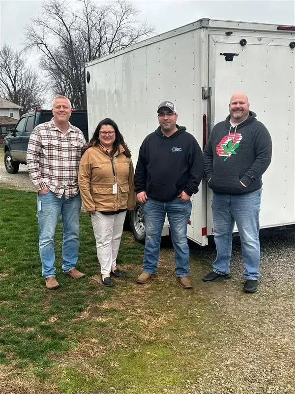 Four people standing outdoors in front of a white trailer on a grassy day