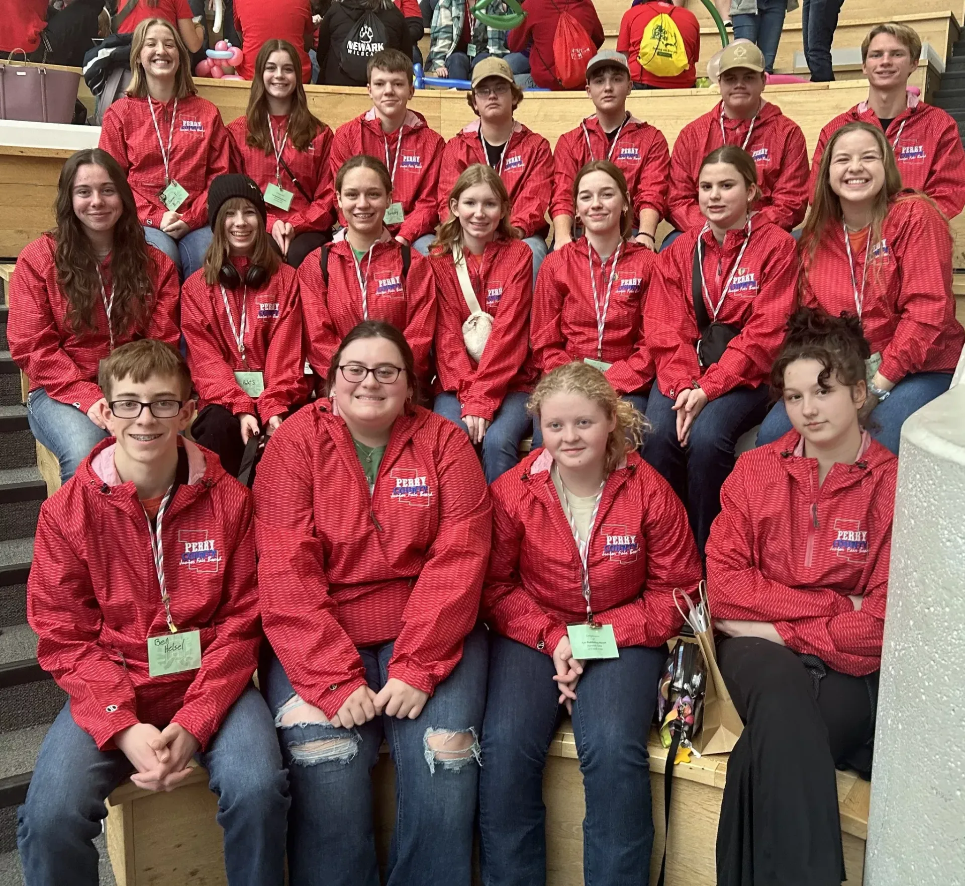 Group of people in red jackets posing on stadium bleachers, smiling at the camera.
