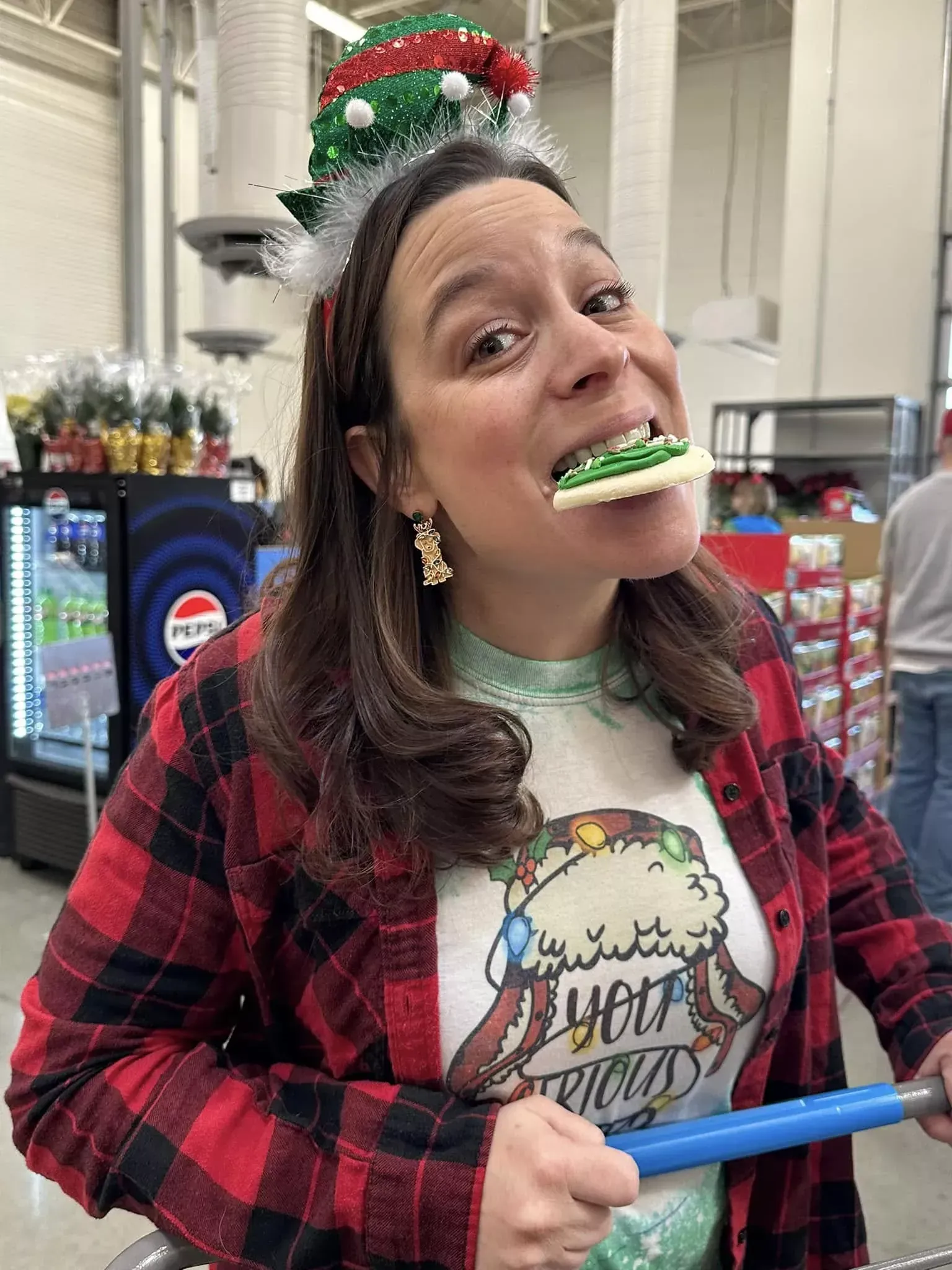 Woman in red plaid shirt and festive hat bites a green and white treat in a store aisle.