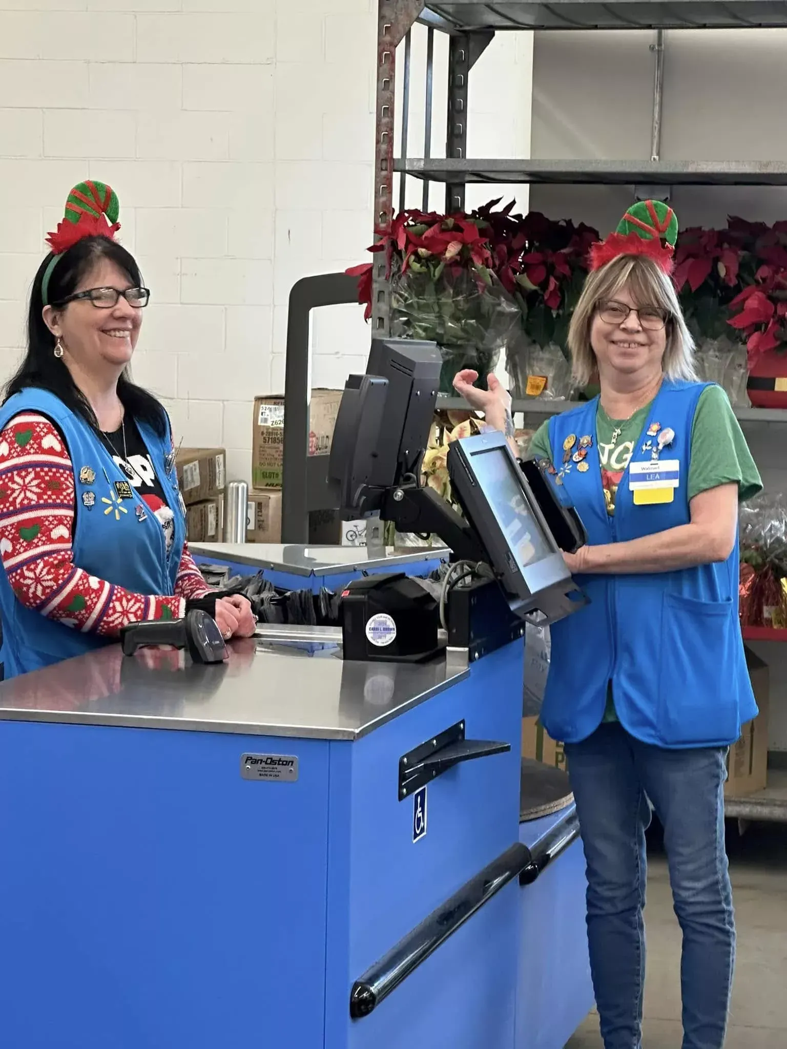 Two women in blue vests stand beside a blue machine in a warehouse, one smiling and waving.