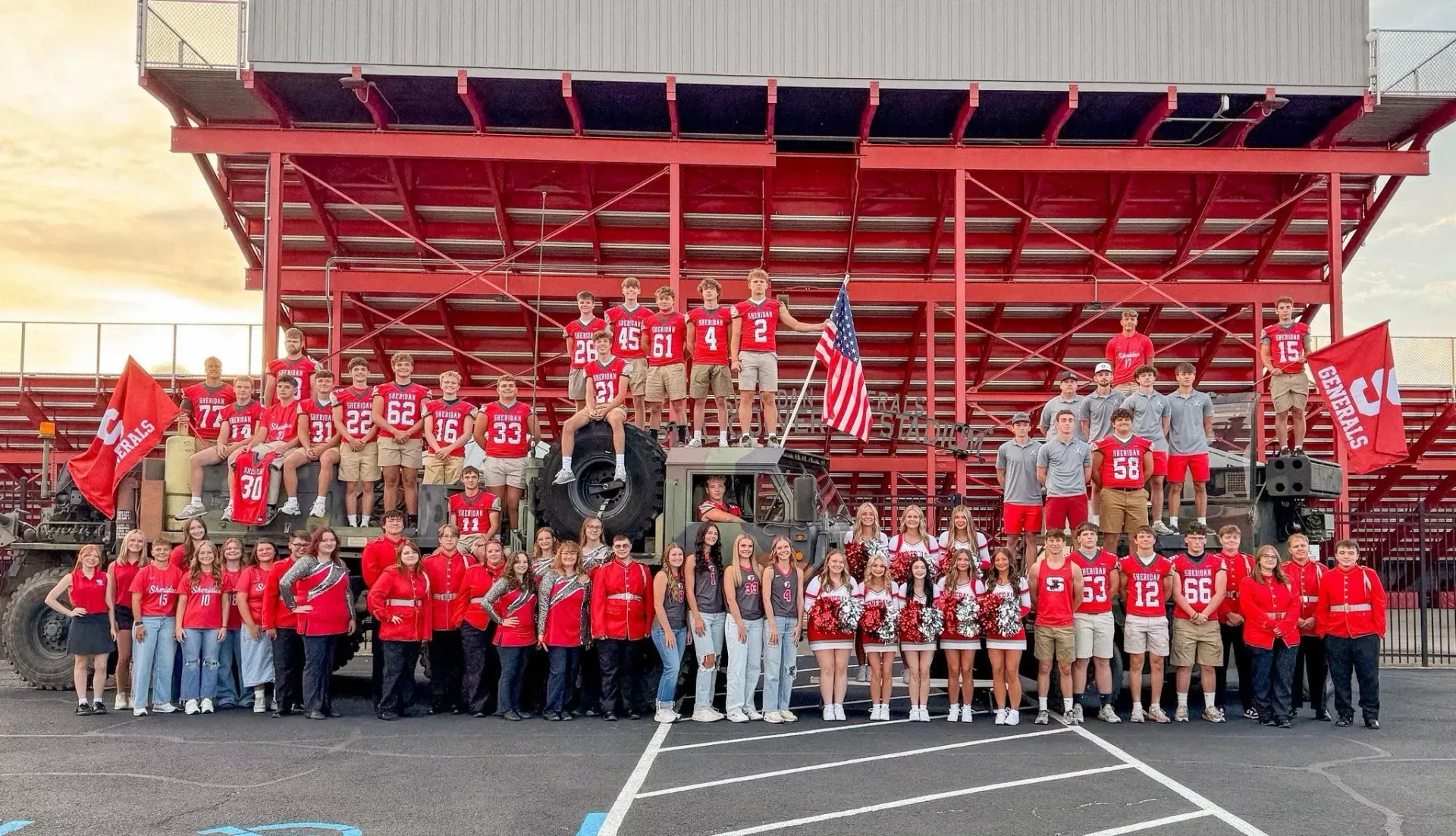 Large group in red shirts posing on bleachers at a sports field, with a red pavilion and flags behind them