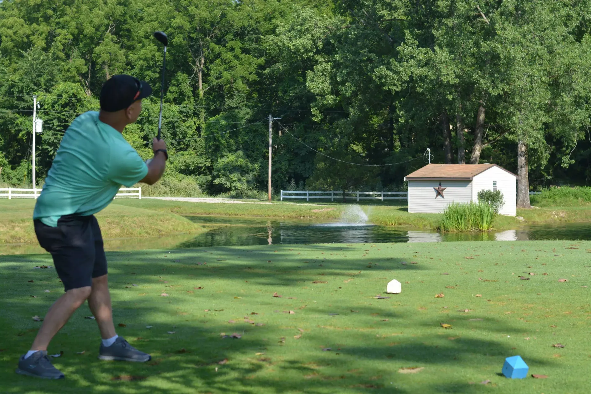 Golfer in teal shirt swings at a ball on a sunny green course near a pond and small white building
